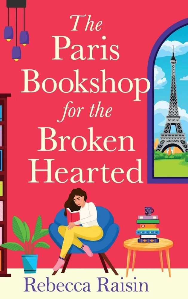 "The Paris Bookshop for the Broken Hearted" by Rebecca Raisin. A woman reading in a blue armchair against a bright pink background. The cozy scene includes a stack of books, a potted plant, and a large arched window with a view of the Eiffel Tower.