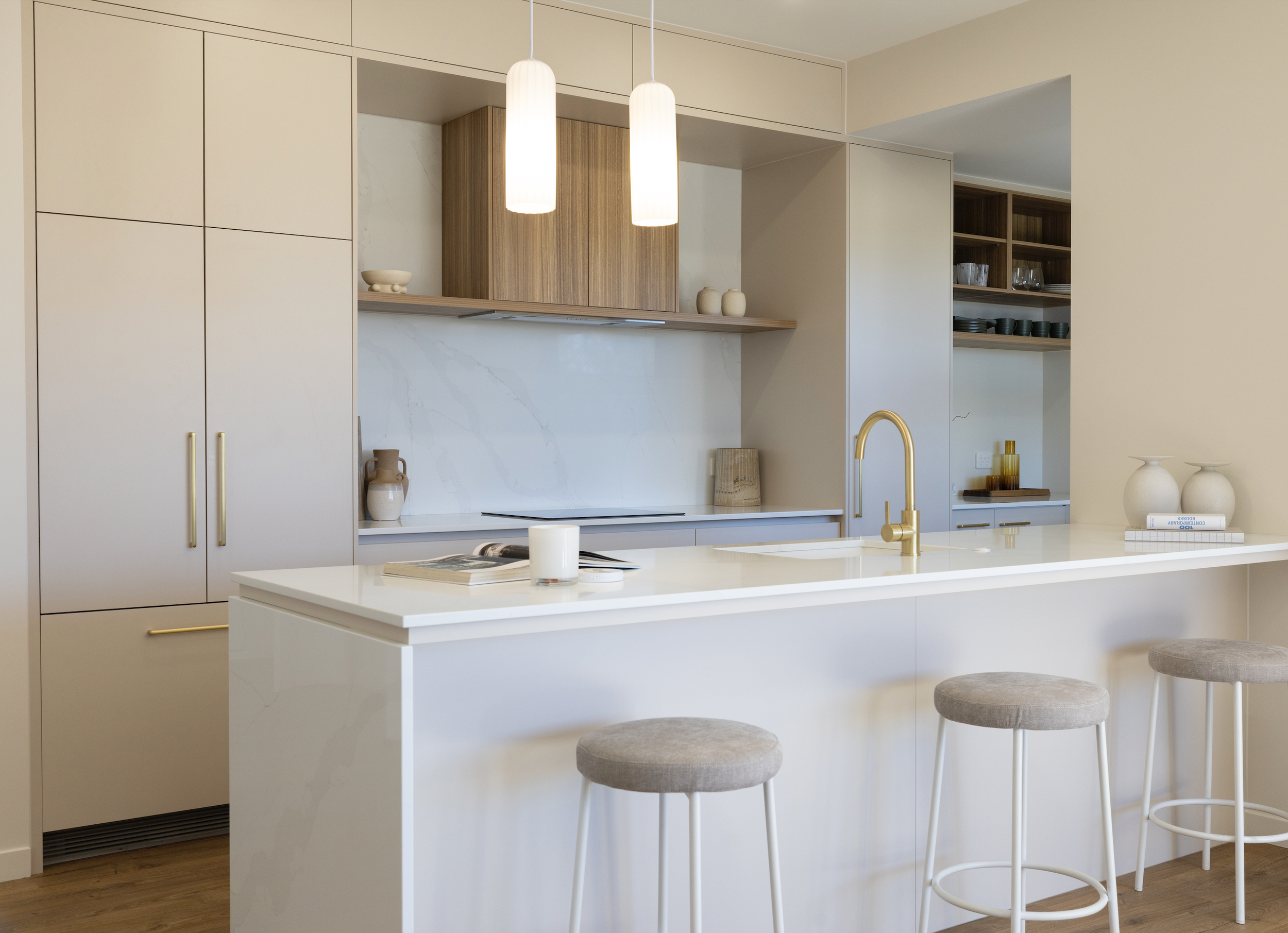Modern kitchen with white island counter, beige cabinetry, open wooden shelves with dishes, and three pendant lights above the island. Allure Interiors design.
