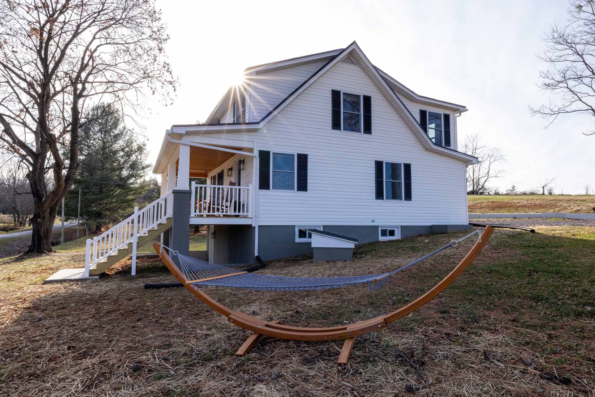 A white house with black shutters and a front porch with stairs, set in a yard with a hammock, surrounded by leafless trees and an open sky.