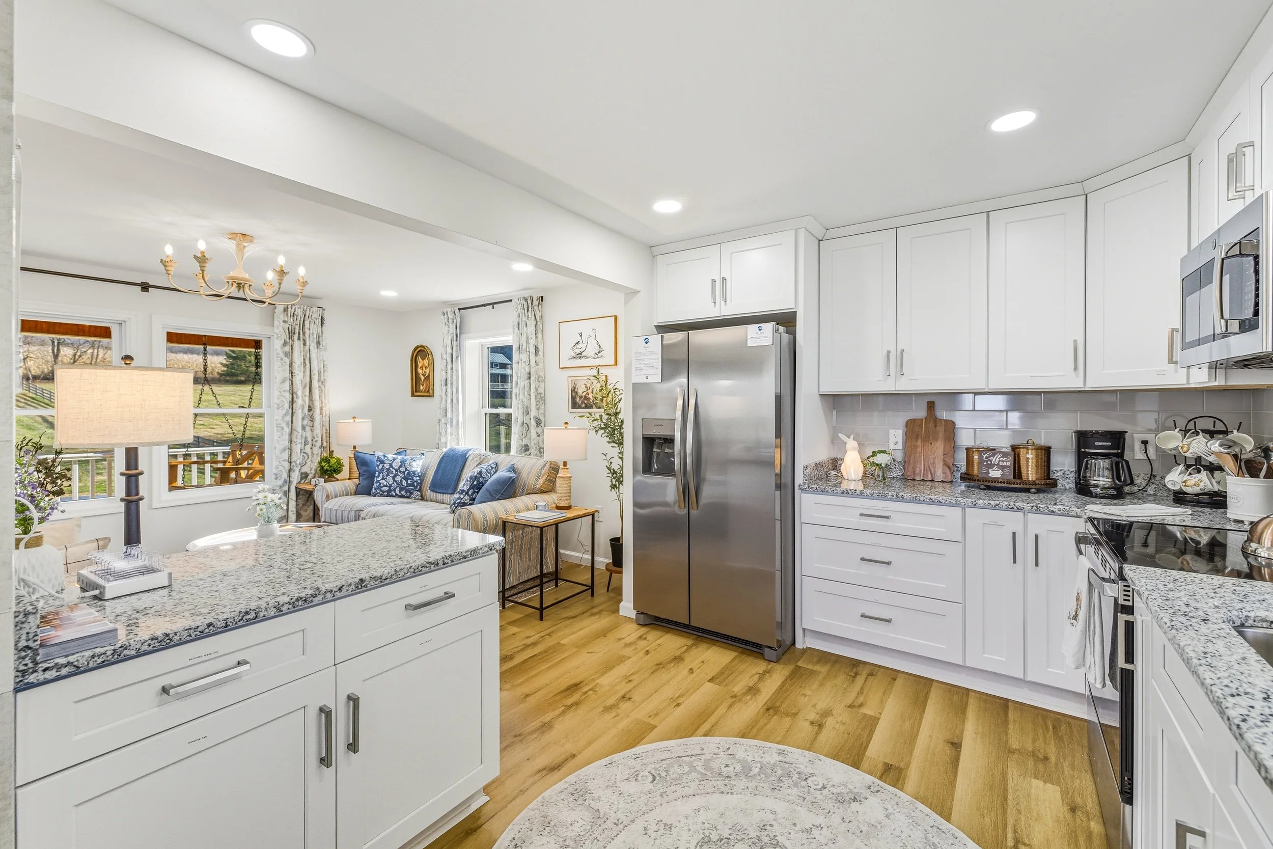 A bright kitchen with white cabinets, stainless steel refrigerator, and granite countertops. The open plan extends to a cozy living area with a sofa, lamps, and decorative wall art. Large windows with curtains let in natural light.
