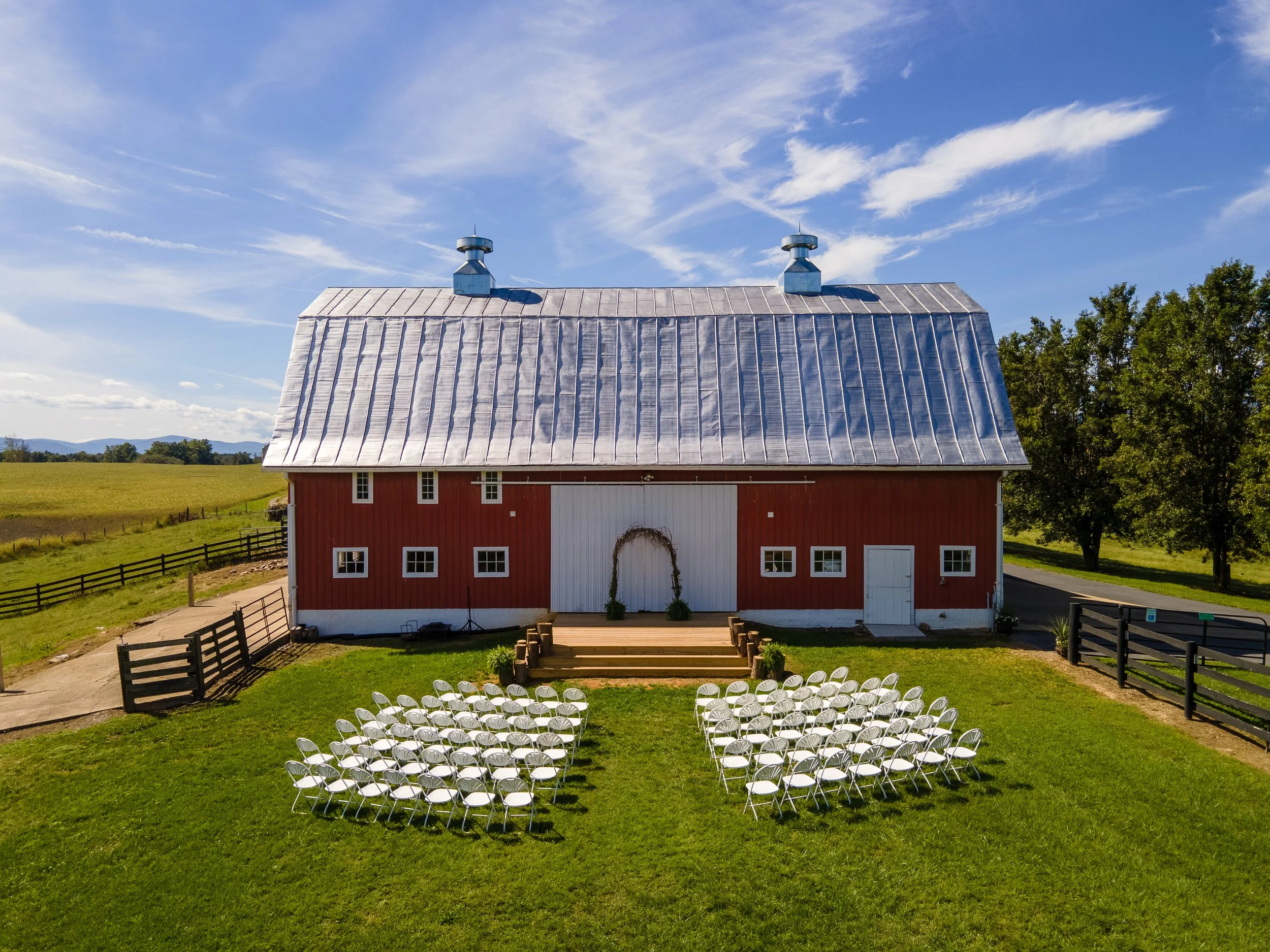 Outdoor wedding setup in front of a red barn with a metallic roof, featuring rows of white folding chairs on a green lawn under a blue sky.