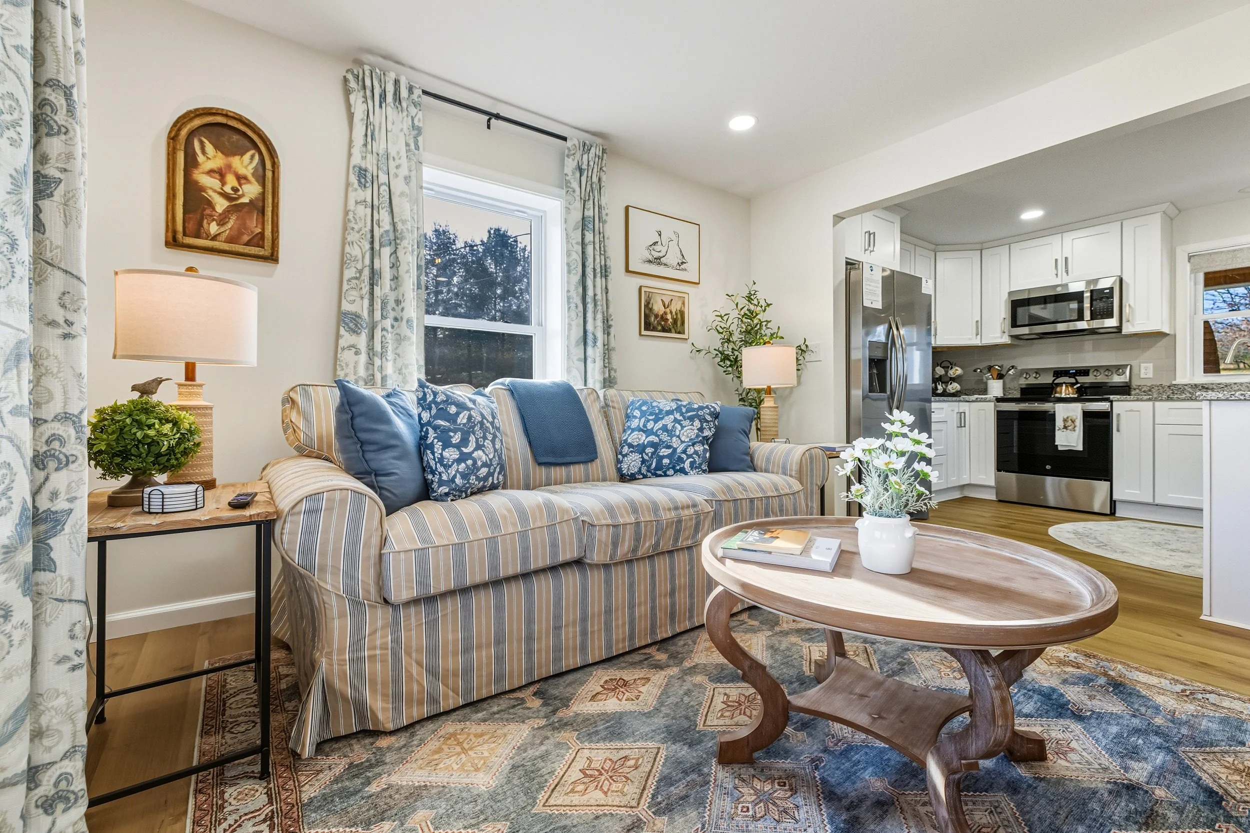 Living room with a striped sofa, blue cushions, a wooden coffee table with books, a side table with a lamp and plant, and a view of the kitchen with white cabinets and stainless steel appliances.