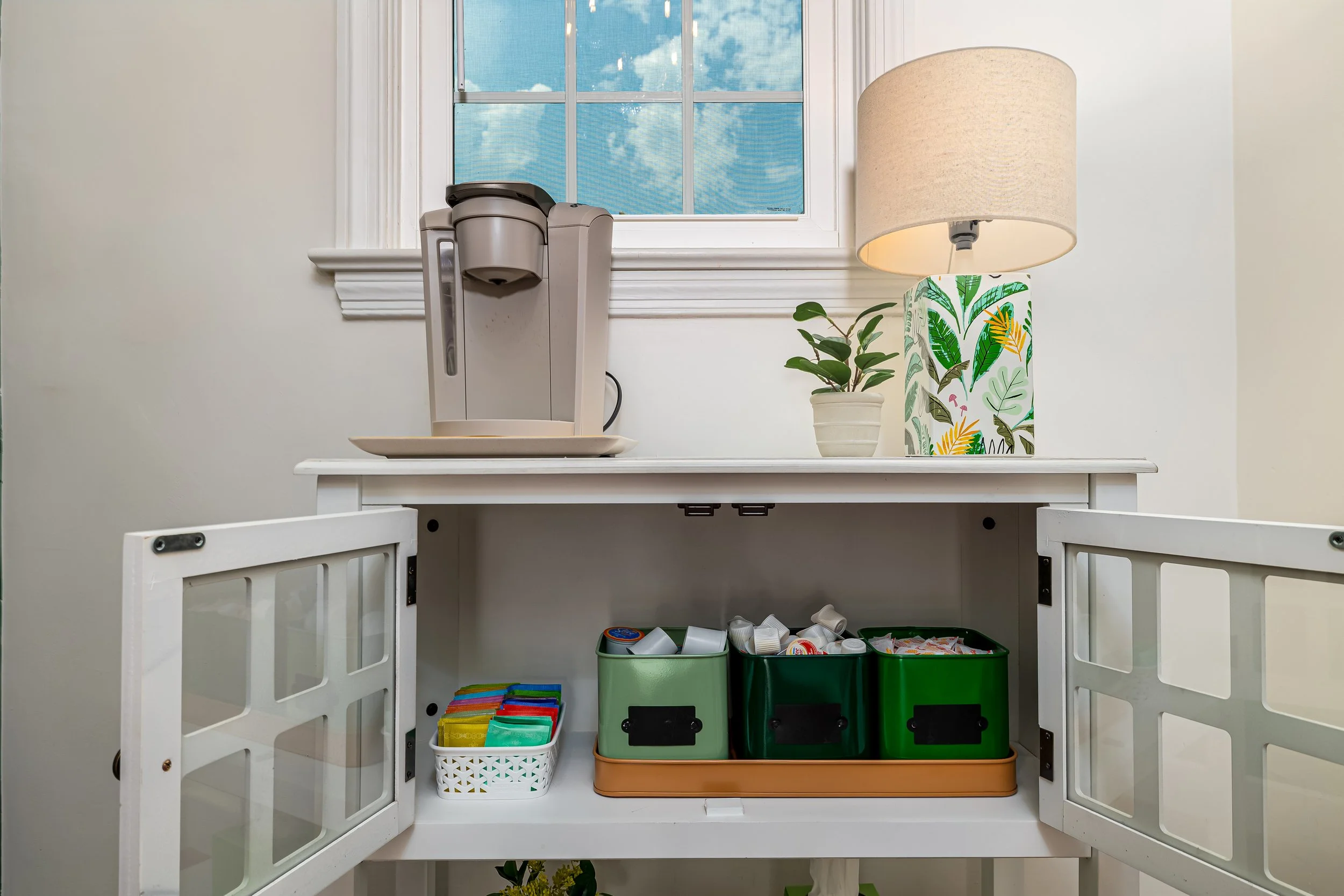 Open kitchen cabinet with a coffee maker on top, a potted plant, and a decorative lamp with a leafy pattern. The cabinet contains labeled containers and cleaning supplies inside.