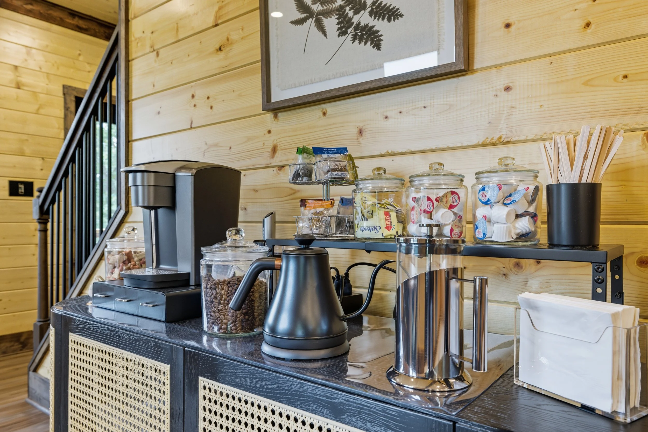 A coffee station with a black countertop, a coffee machine, jars of coffee and sugar, and a container with wooden stir sticks, set against a light wood-paneled wall with a framed botanical print.
