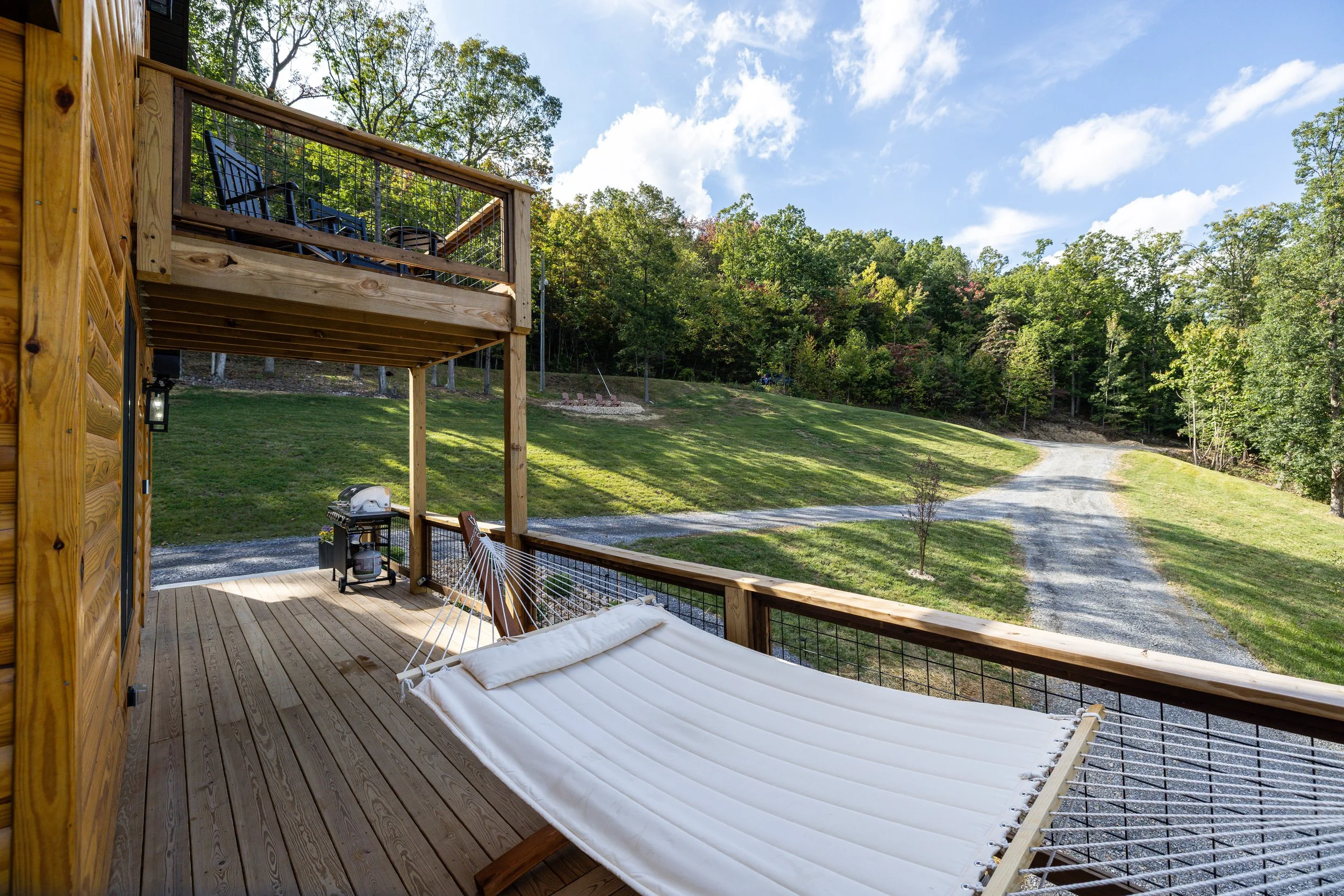 A wooden deck with outdoor furniture, including a white cushioned lounge chair, overlooking a grassy yard and wooded hillside under a blue sky with some clouds.