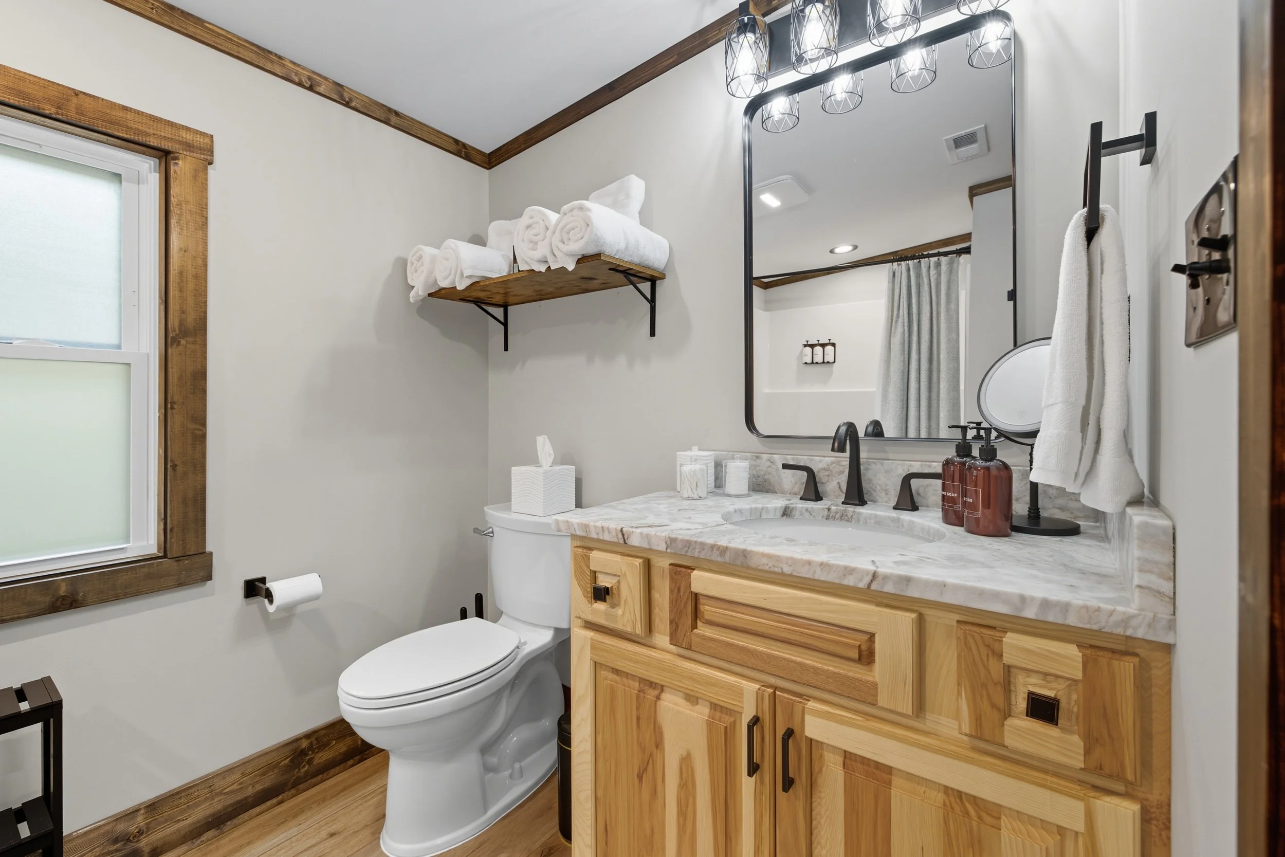 A bathroom with a marble countertop vanity, a mirror, and wall-mounted lighting. Contains a toilet with a tissue box, towels on a wall shelf, and a window with frosted glass and wood trim.