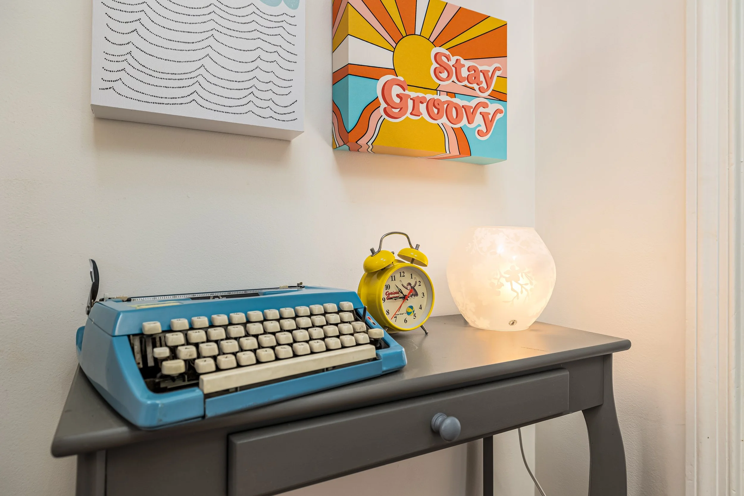 A desk with a vintage blue typewriter, a yellow alarm clock, a decorative lamp, and two wall art pieces, one featuring a sun and colorful lines with the phrase "Stay Groovy."