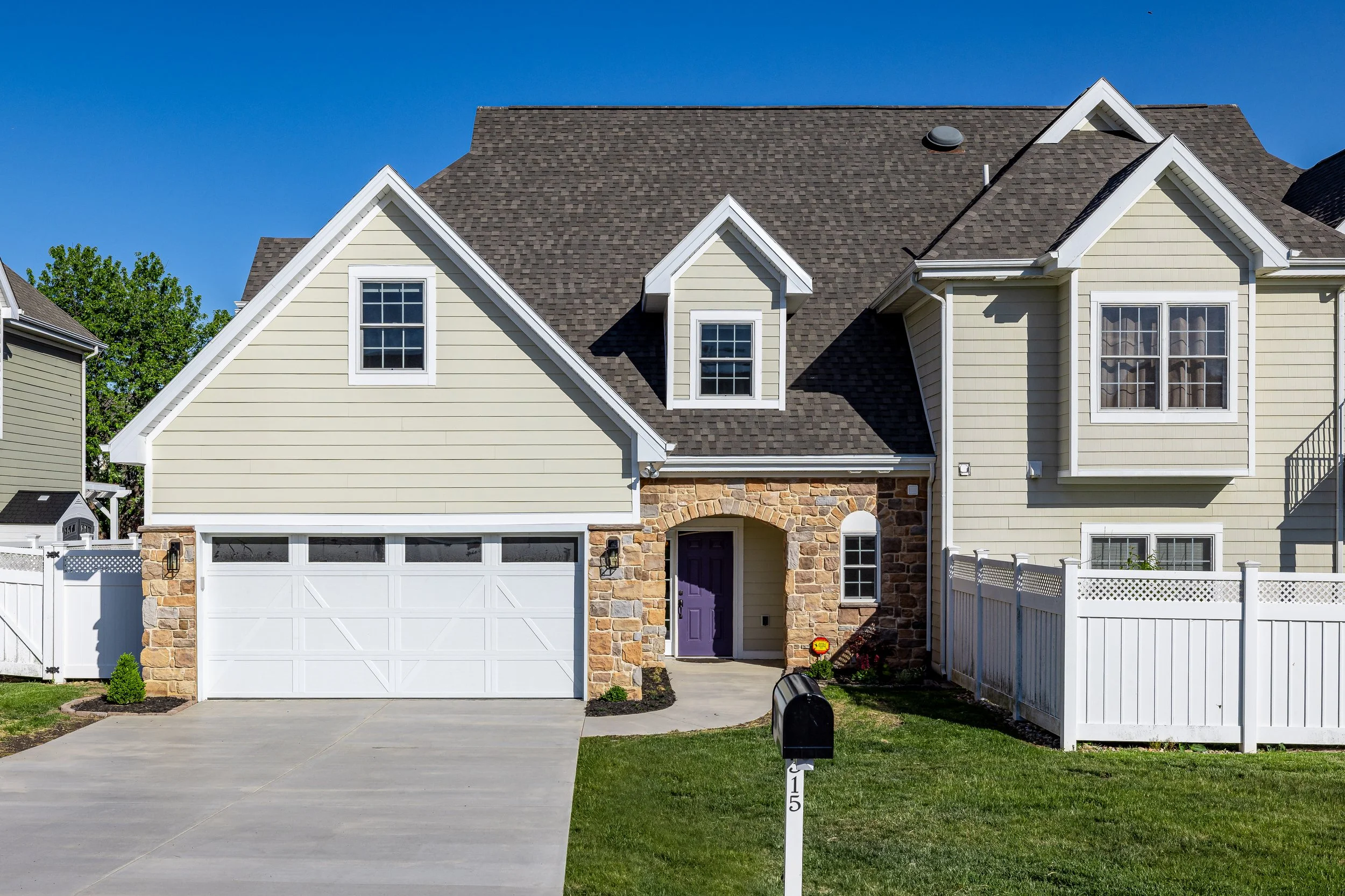 View of a two-story house with beige siding, large roof, and a purple front door. The house has a white garage door and a stone archway above the front door. There is a white fence on the right side, a mailbox in the front yard, and a green lawn with