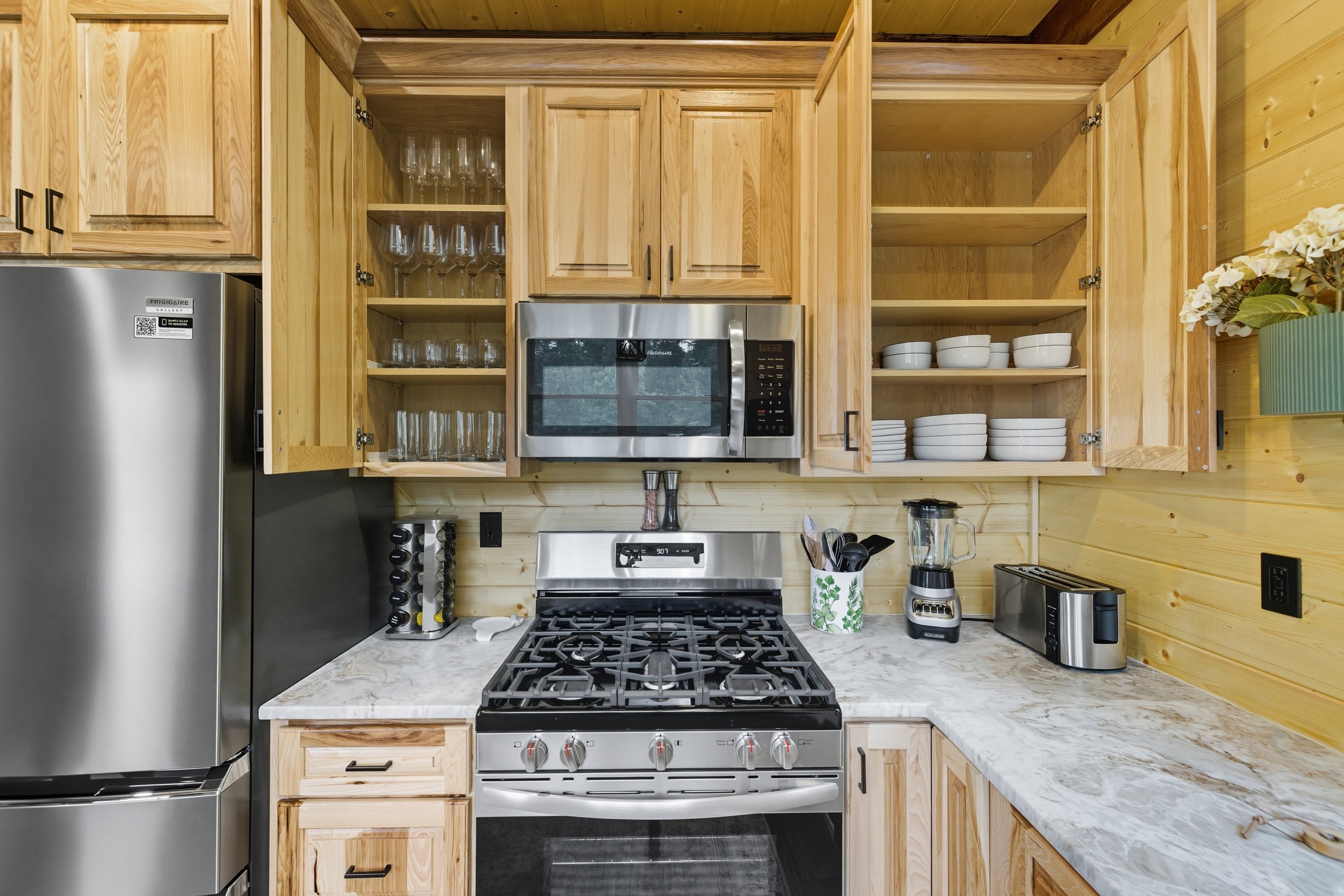 Kitchen with light wood cabinets, marble countertop, stainless steel refrigerator, microwave with glassware inside, stove, toaster, and a blender, with a small potted plant of white flowers on the right.