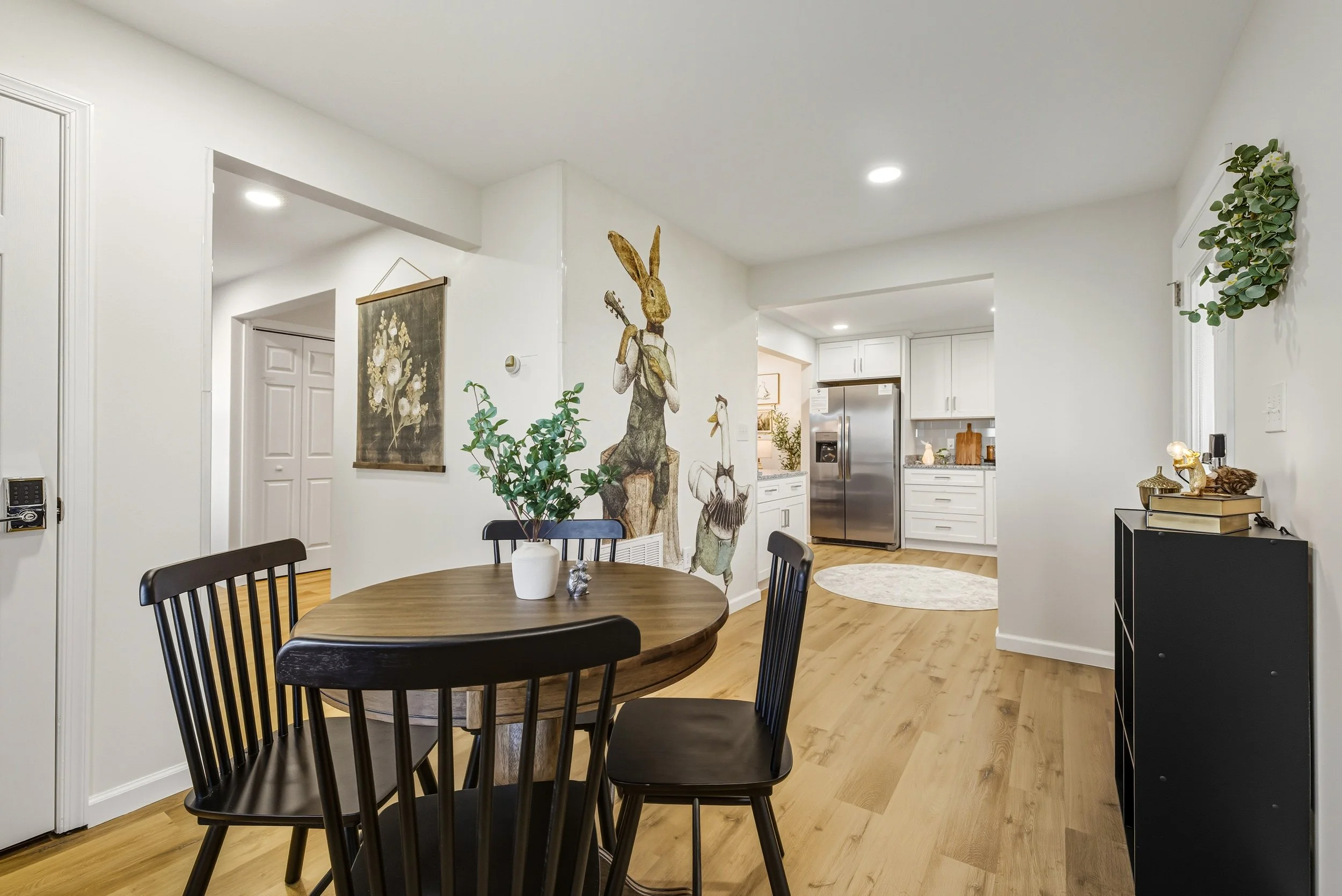 Open concept living and dining area with white walls, hardwood floors, a round dining table with black chairs, green plants, wall art, and a kitchen in the background with white cabinets and stainless steel refrigerator.