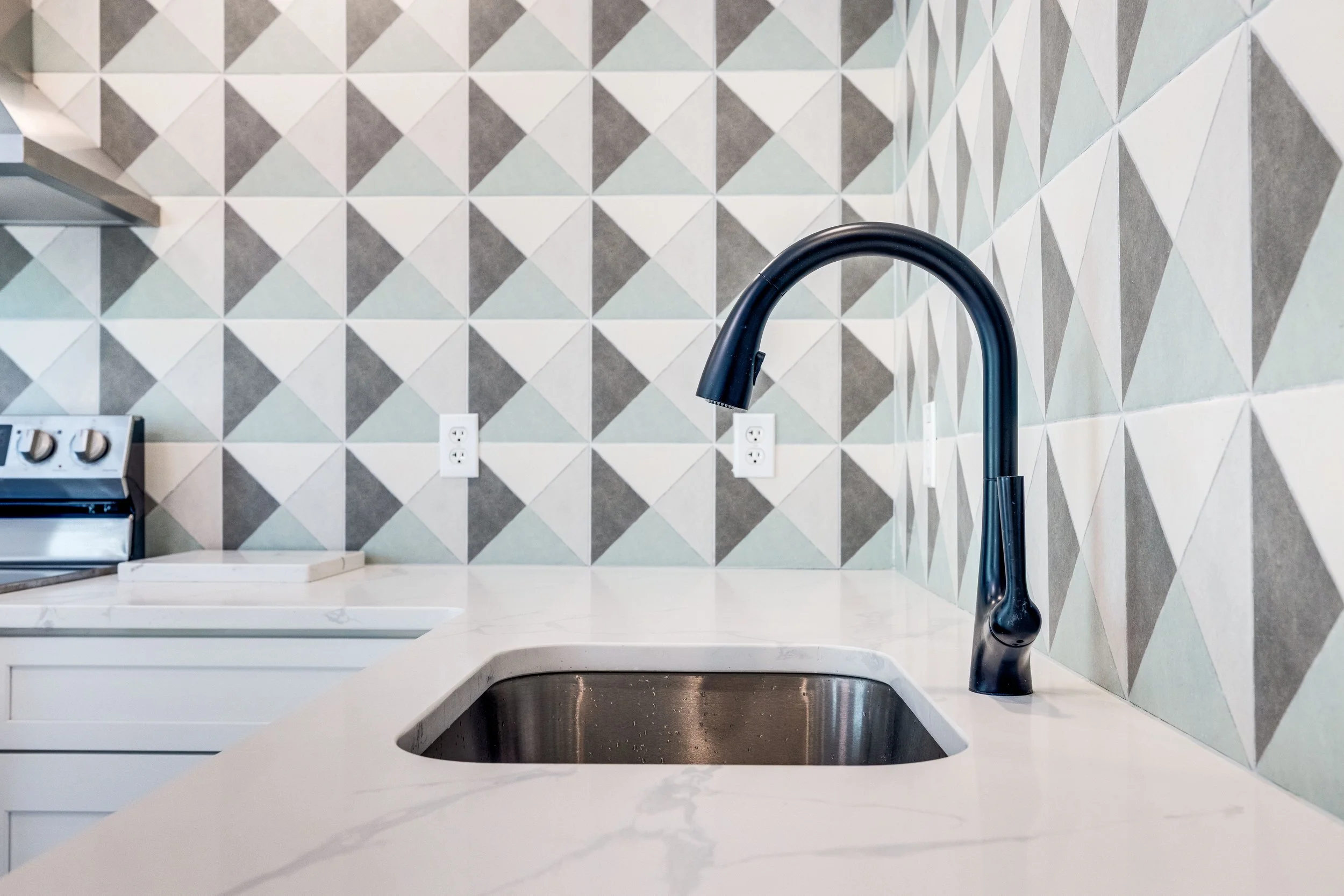 A kitchen countertop with a black faucet, a white cutting board, and a stove, against a wall with geometric patterned tiles in shades of gray, white, and light blue.