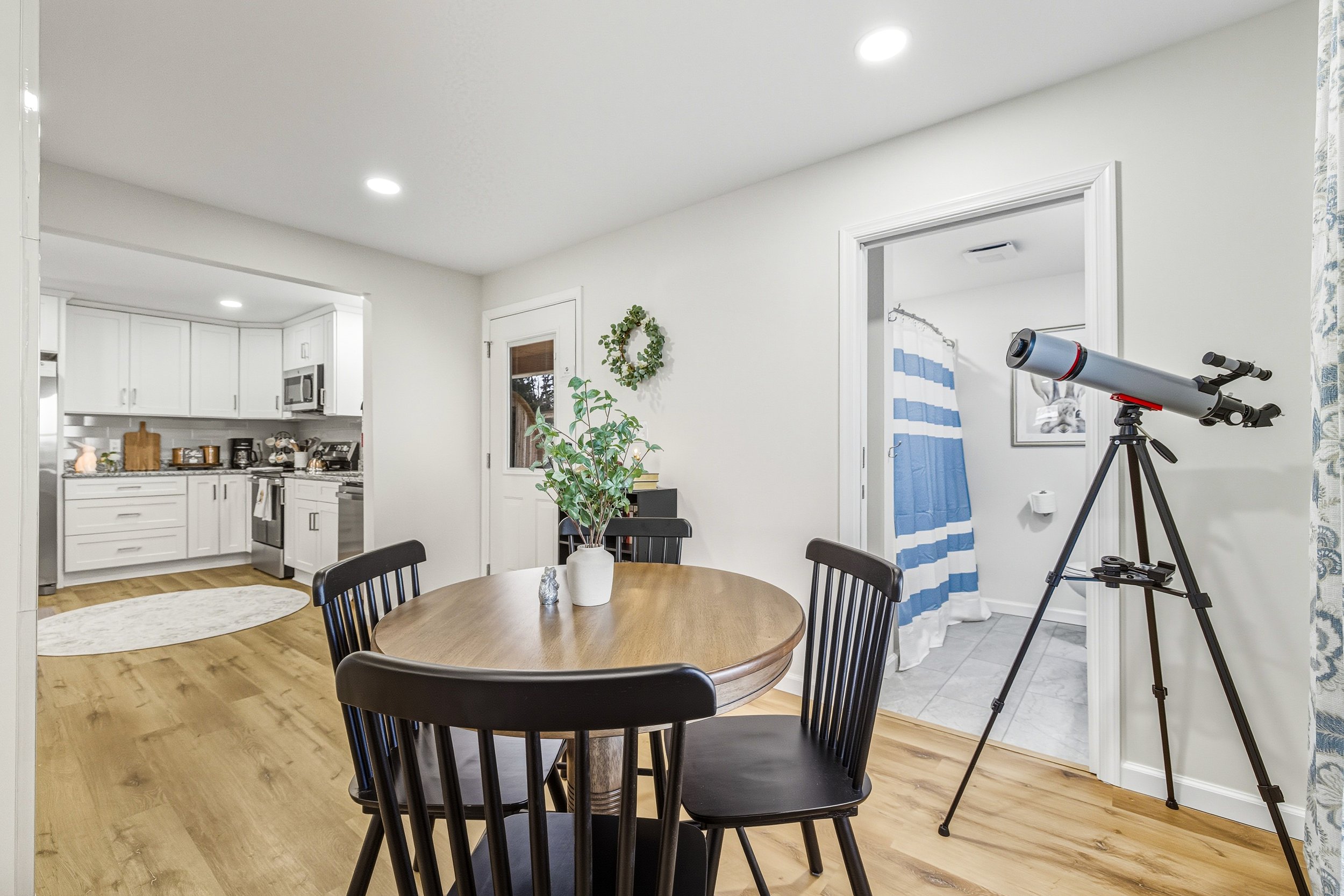A dining area with a round wooden table and black chairs, a potted plant, and a telescope pointed towards the window. The background shows a kitchen with white cabinets and appliances, and a doorway leading to a bathroom with a blue and white shower 