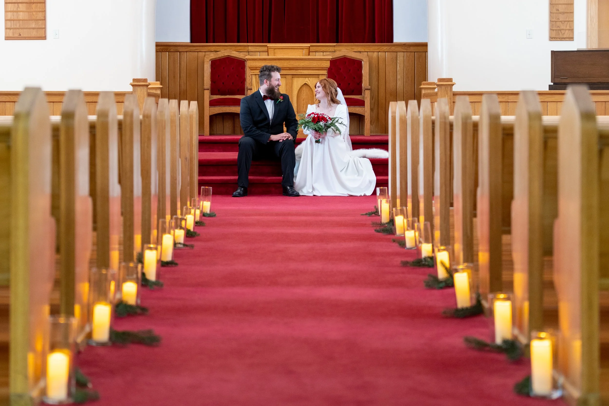 A bride and groom sitting together on the steps of an altar inside a church, surrounded by candles and wooden pews, with a red curtain backdrop.