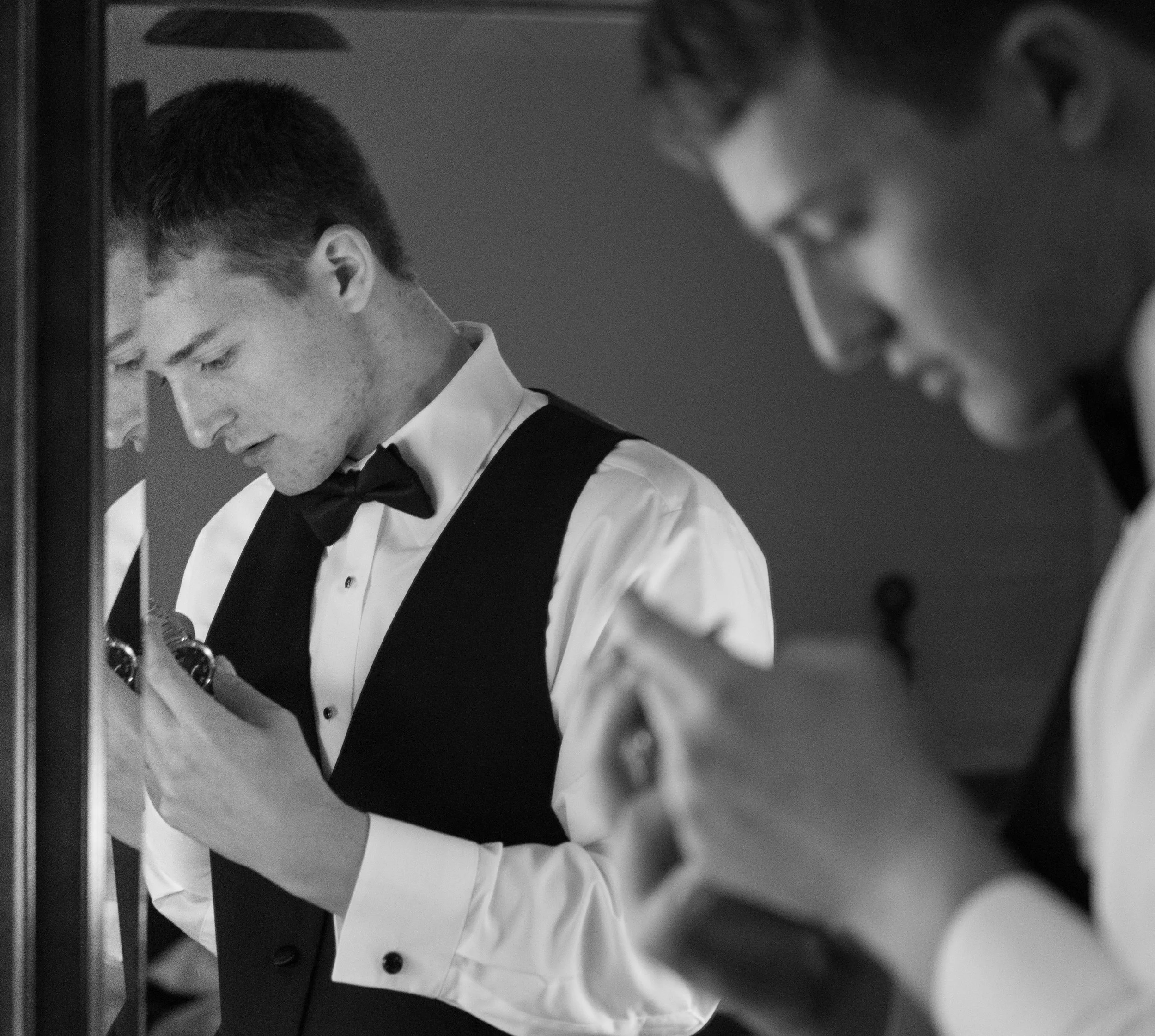 Man in formal attire with bow tie adjusting cufflinks in a mirror.