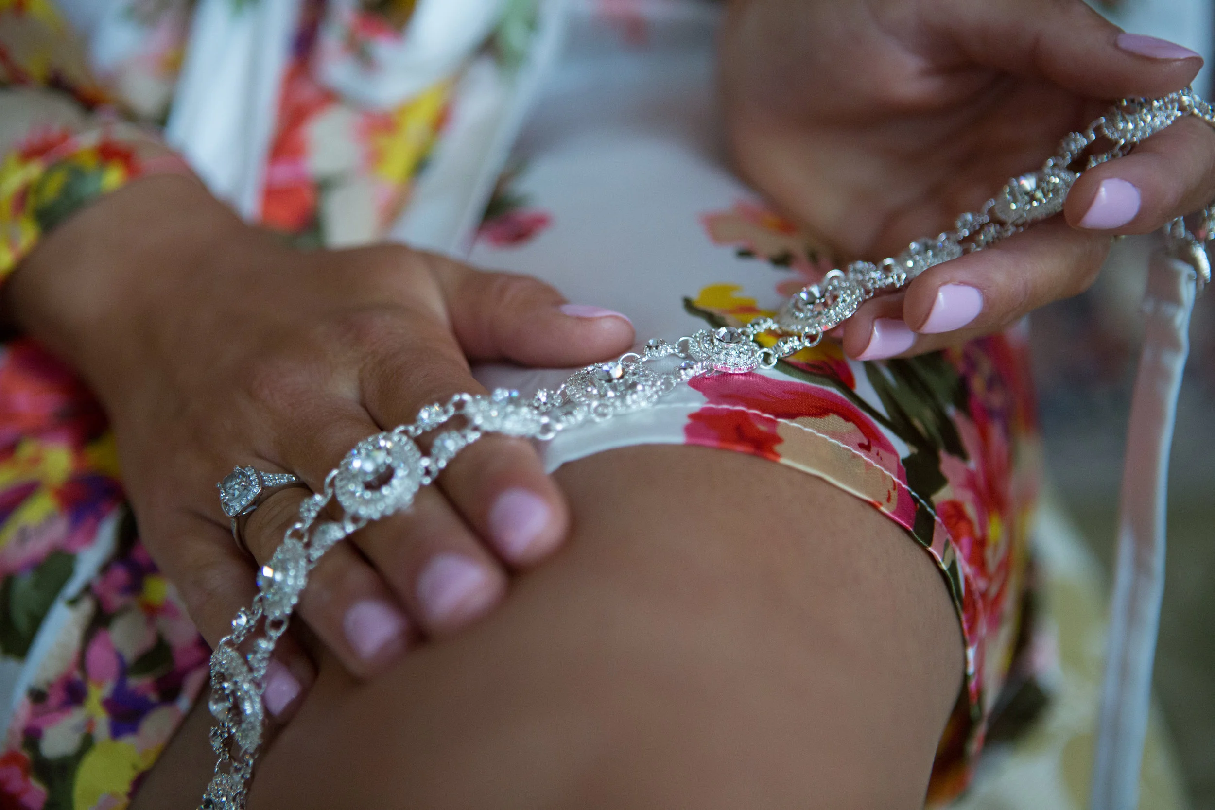 Close-up of a person wearing a floral dress, holding a jeweled garter belt while showcasing a diamond ring on their finger.