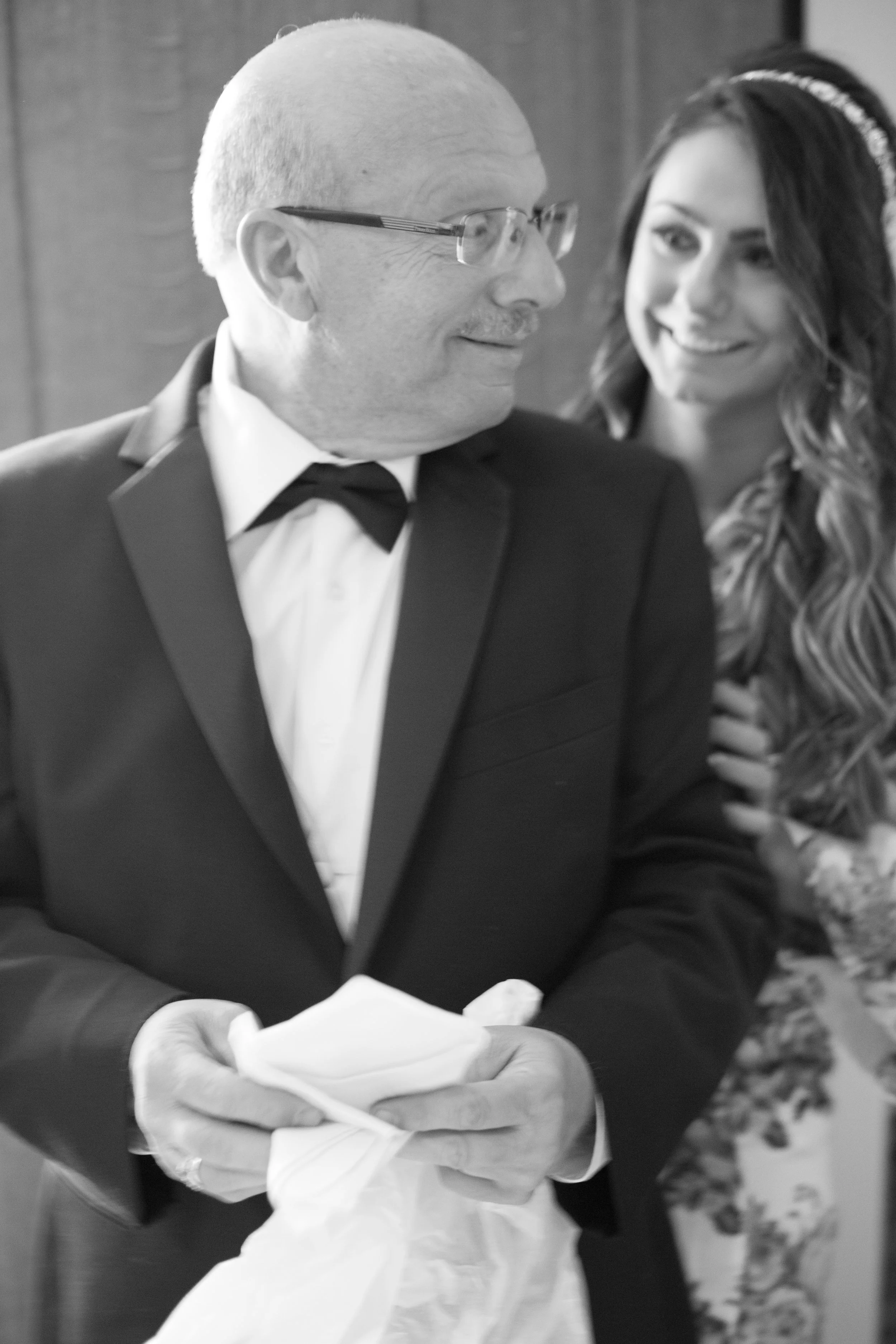 Man in tuxedo with glasses holding a cloth, while a smiling woman is partially visible behind him, indoors.