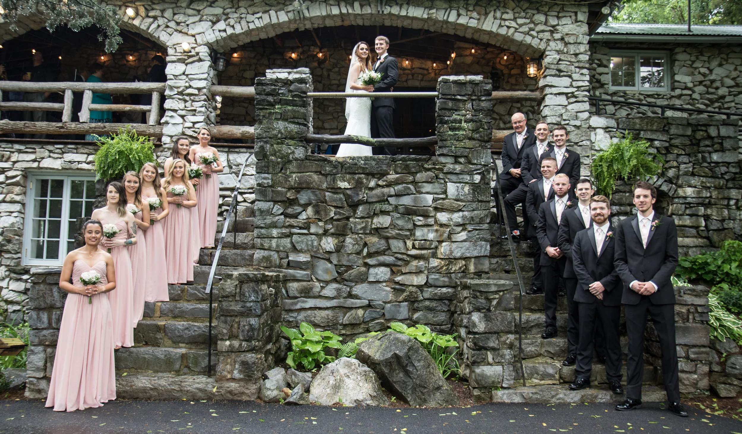 Wedding party with bride, groom, bridesmaids in pink dresses, and groomsmen in black suits posing on stone steps of rustic stone building.