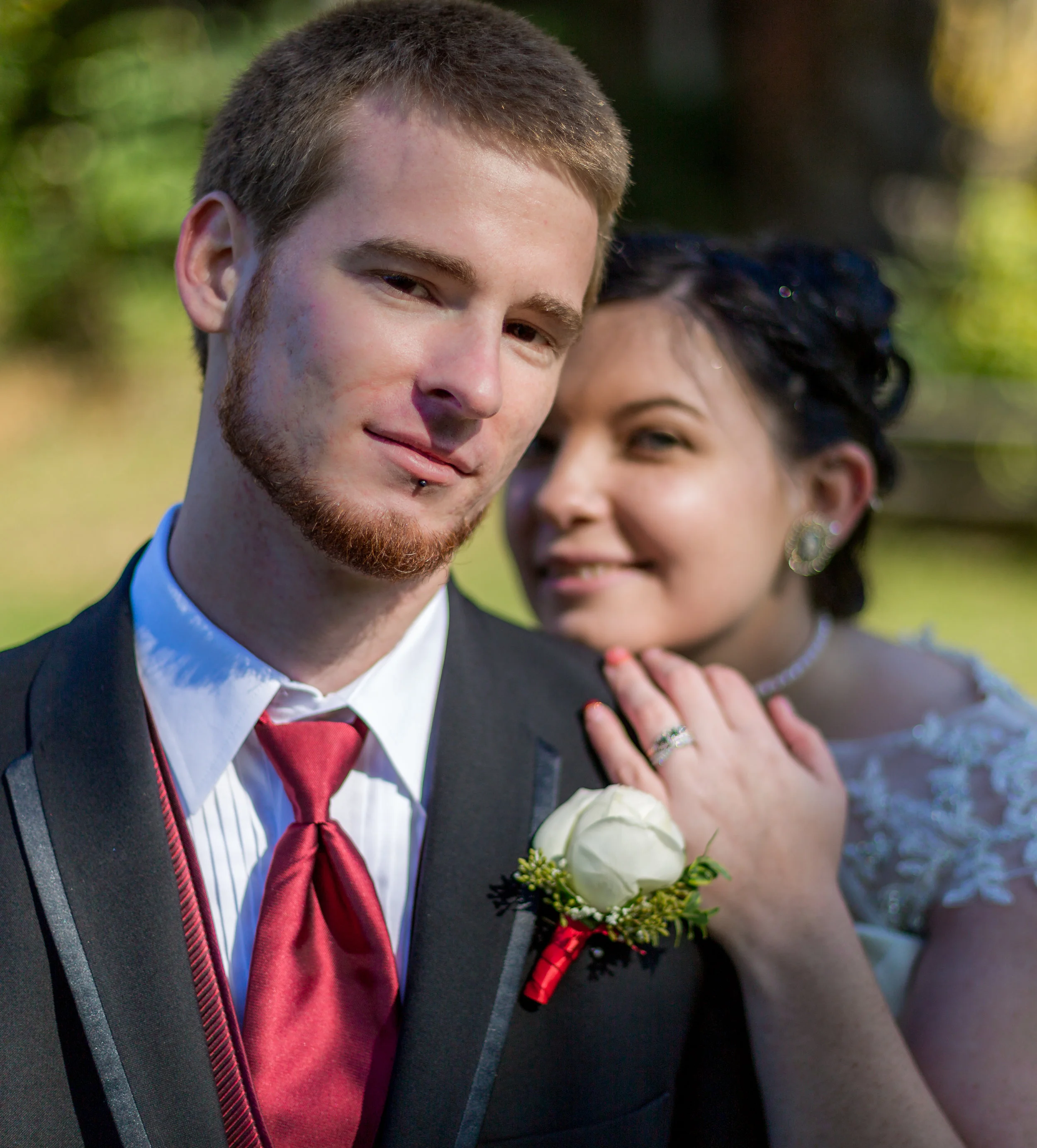 Wedding couple posing, groom in suit with red tie and boutonnière, bride in lace wedding dress, outdoors.