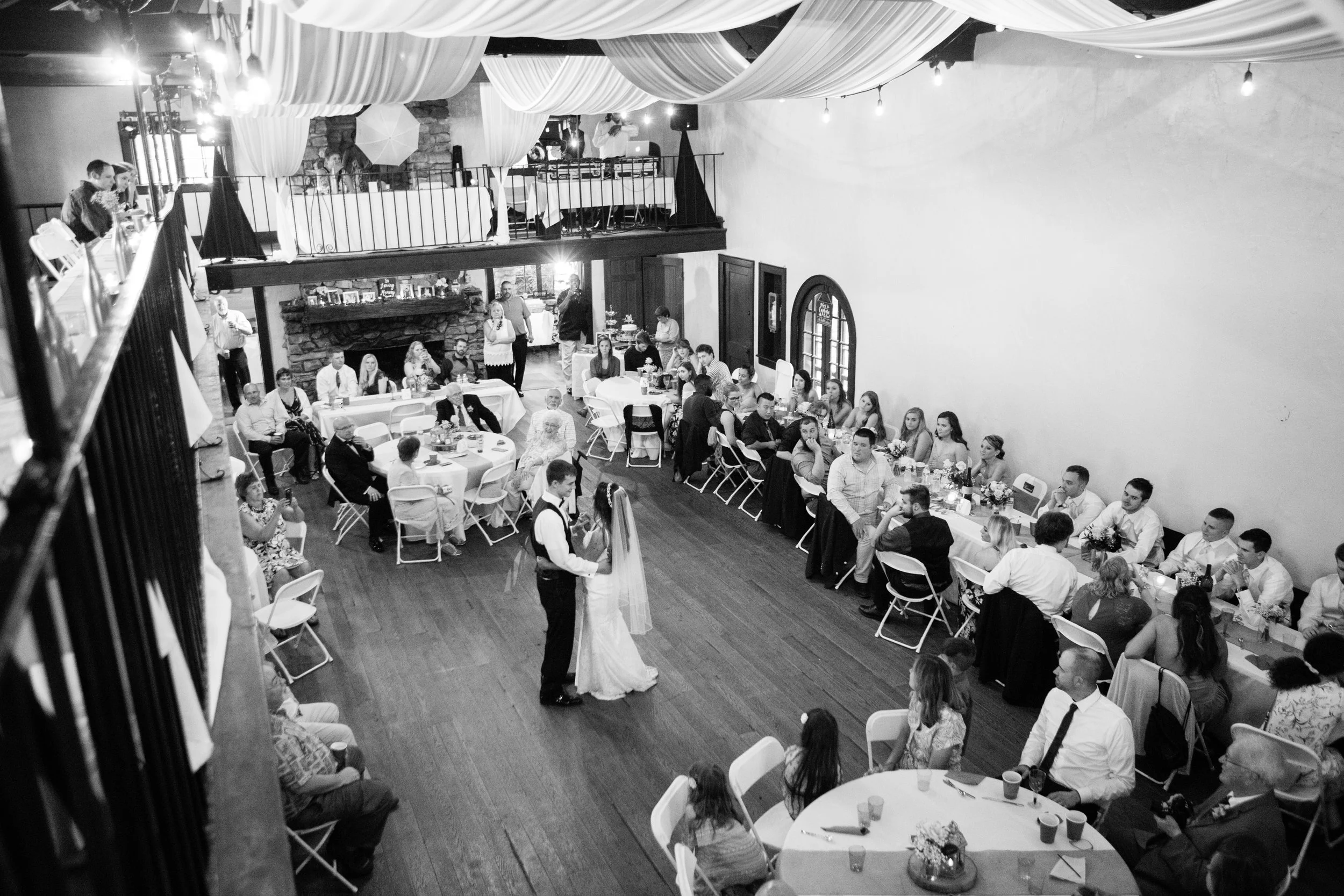 A black and white photo of a wedding reception inside a rustic venue with a high ceiling decorated with draped fabric. Guests are seated at round and rectangular tables, with some standing and others watching a couple in wedding attire dancing on the