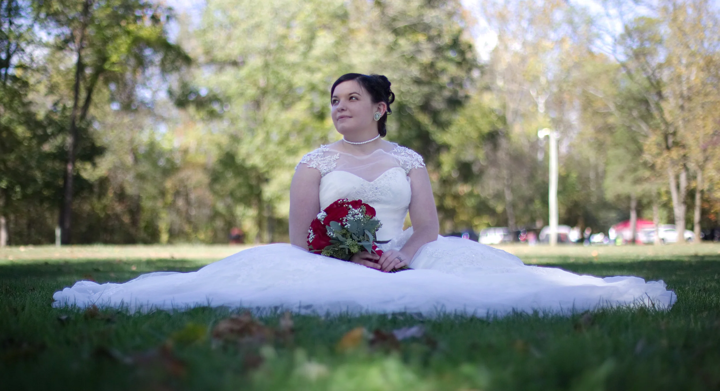 Bride in a white wedding dress sitting on grass, holding a bouquet of red flowers outdoors.