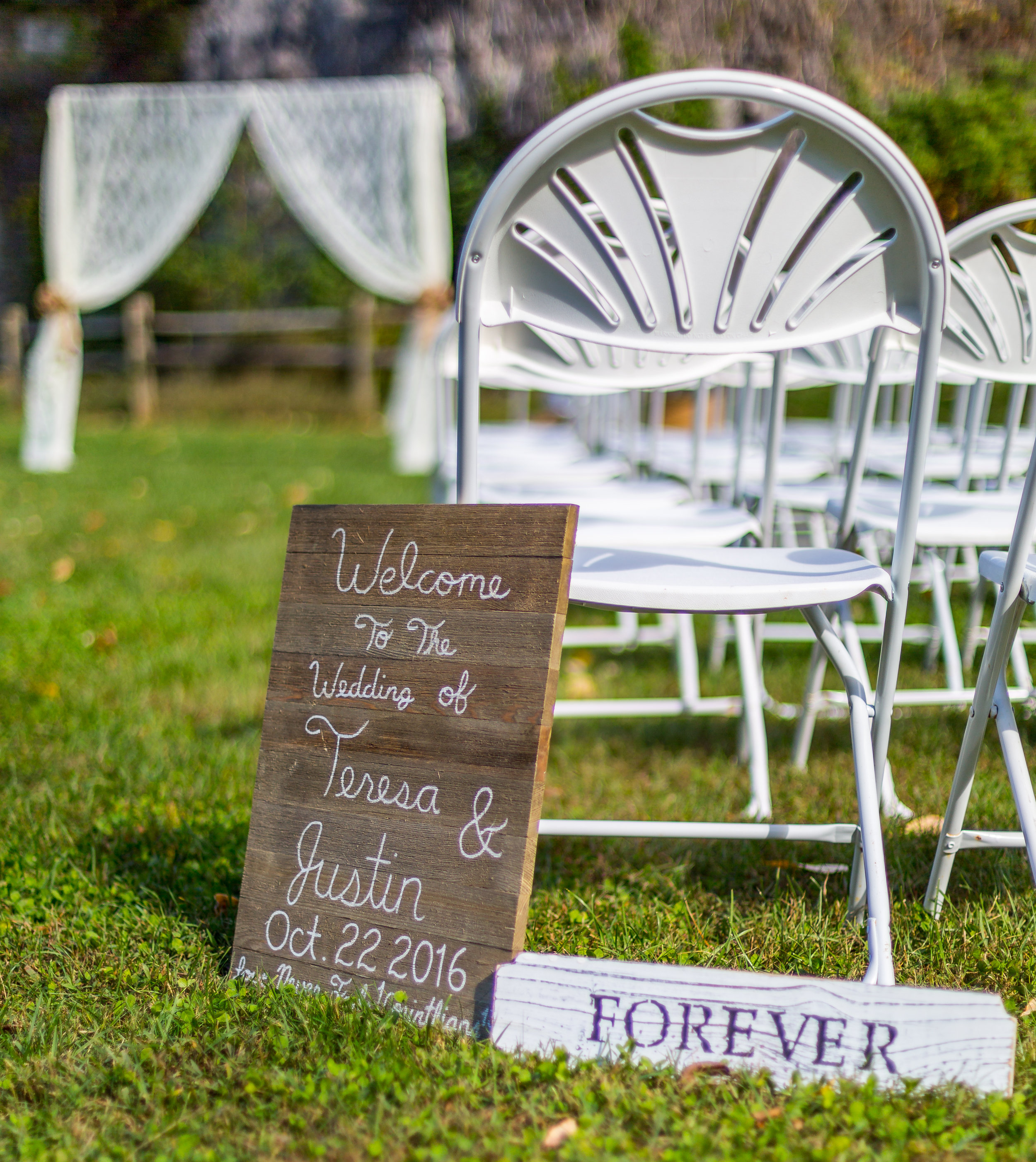 Outdoor wedding setup with white chairs and a wooden welcome sign for Teresa and Justin's wedding on October 22, 2016, with a 'FOREVER' sign on the grass.