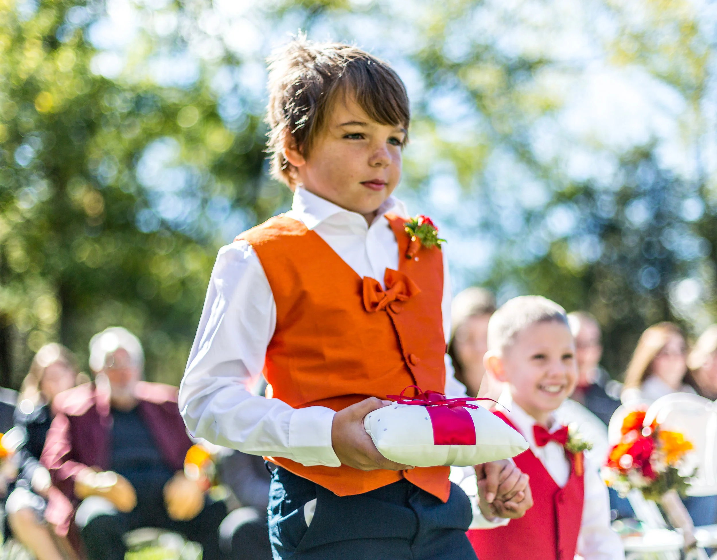 Young boy in orange vest holding a ring bearer pillow at a wedding, smiling child in red vest beside him.