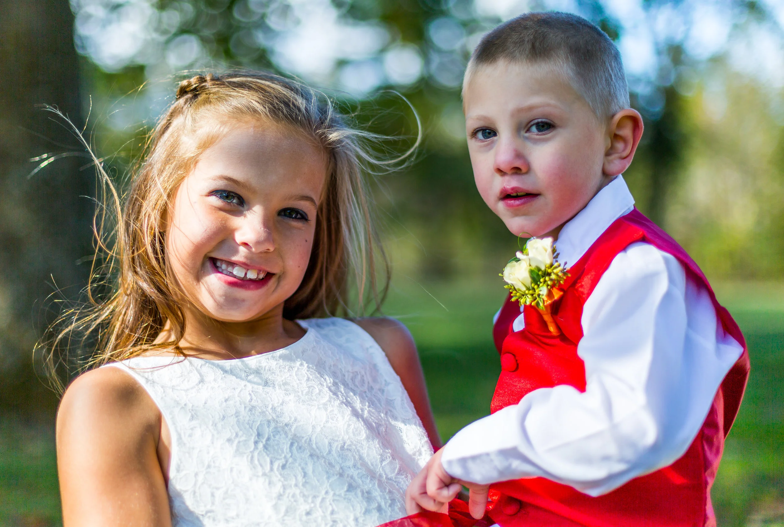 Smiling girl in white dress holding a young boy in a red vest, outdoors in a sunny setting.