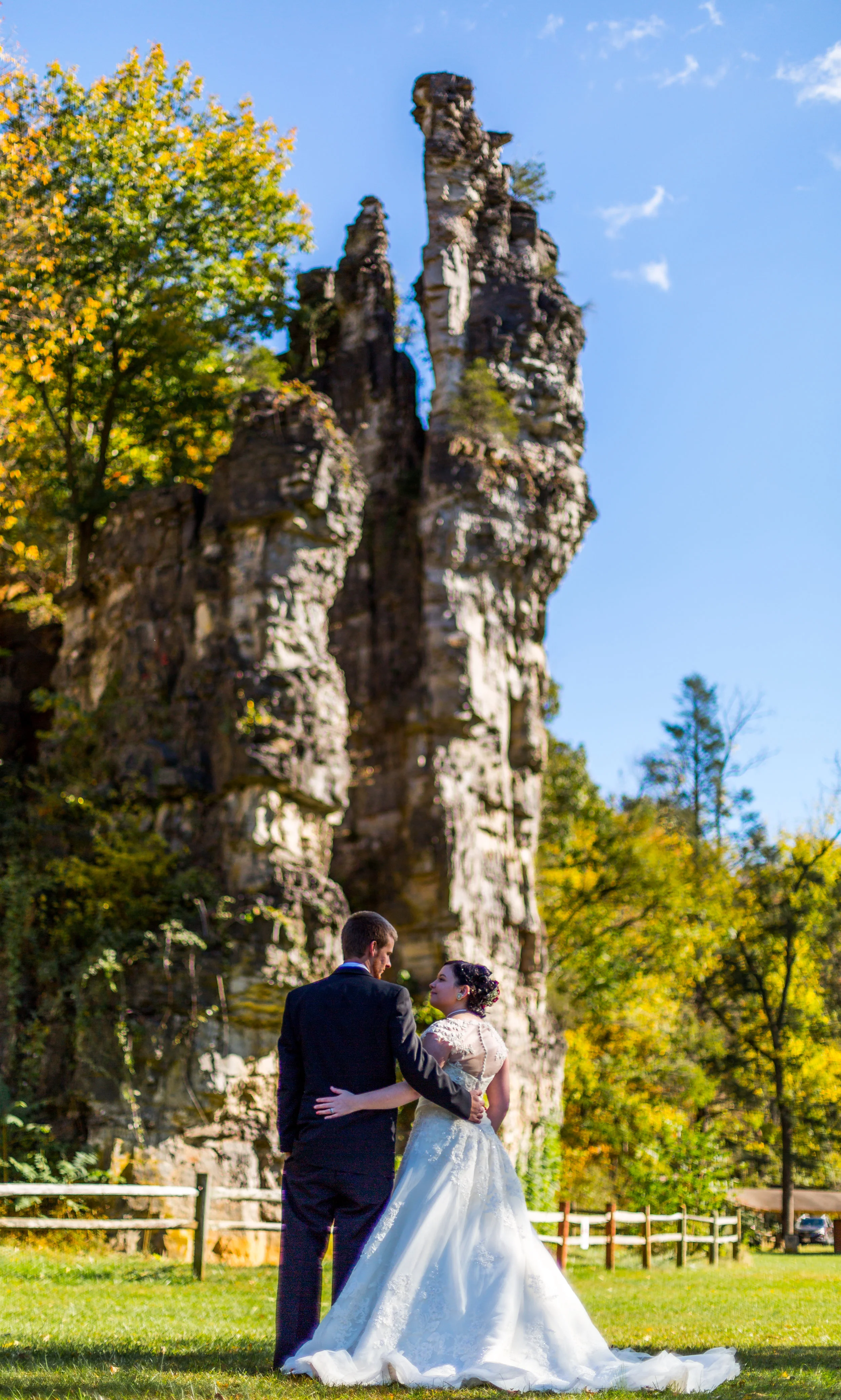 Bride and groom standing in front of rock formations, with autumn trees and blue sky in the background.