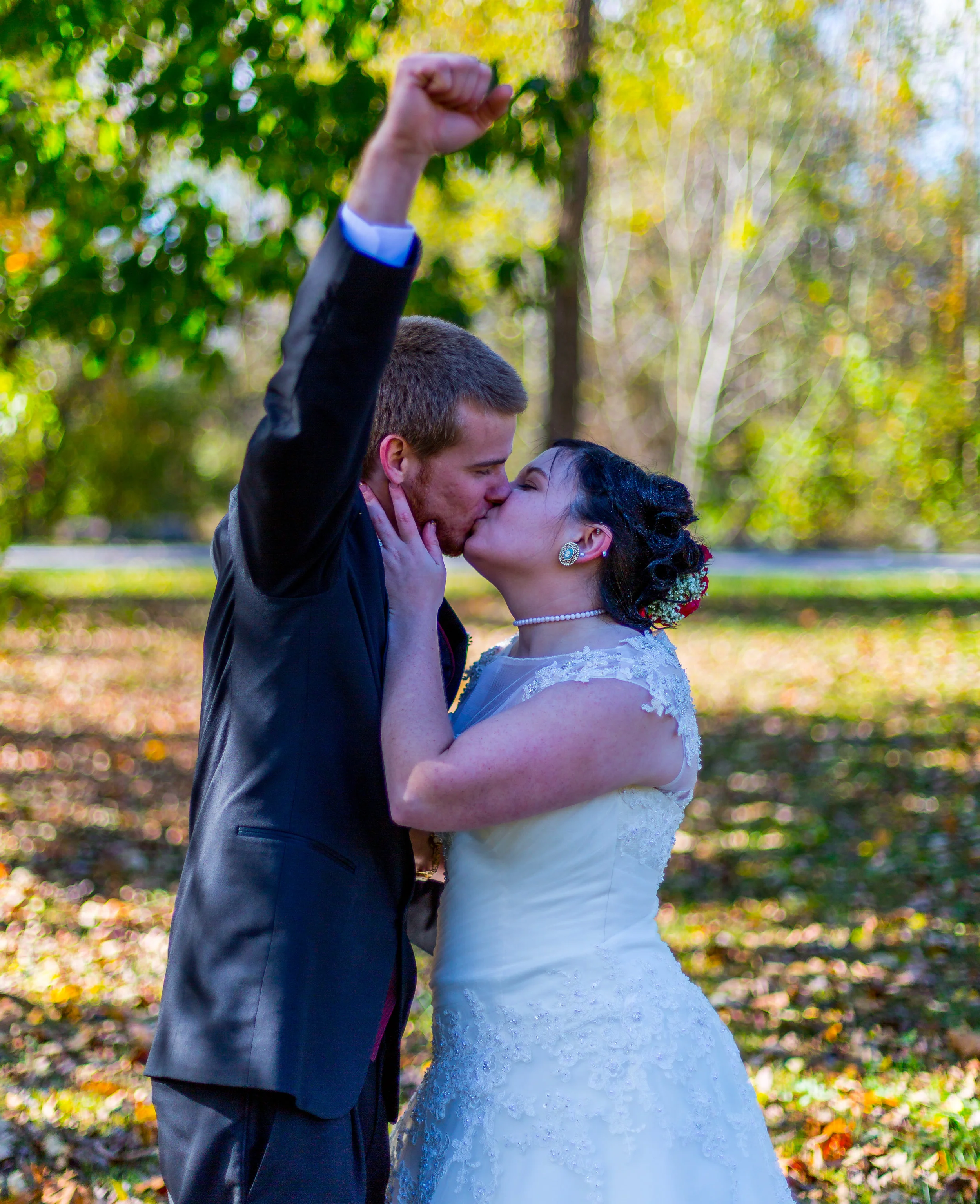 A bride and groom sharing a kiss outdoors during daylight, with the groom raising his fist in celebration. The background features blurred autumn leaves and trees.