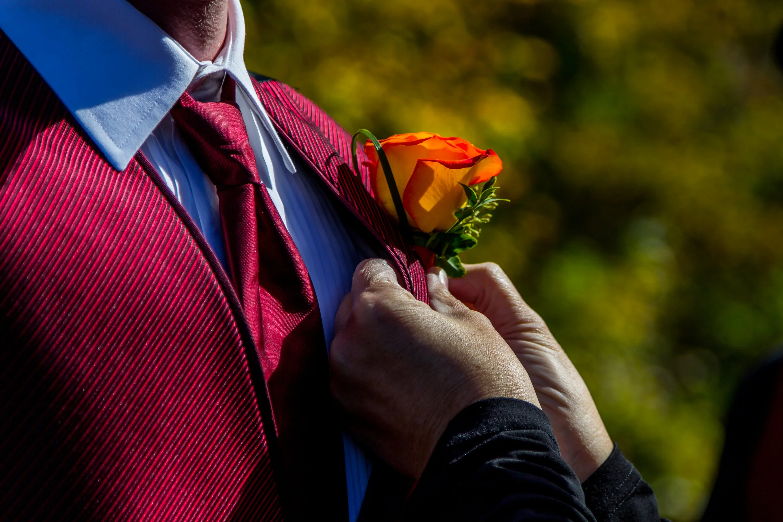 Person wearing a red vest and tie, with another person adjusting an orange rose boutonniere on the lapel.