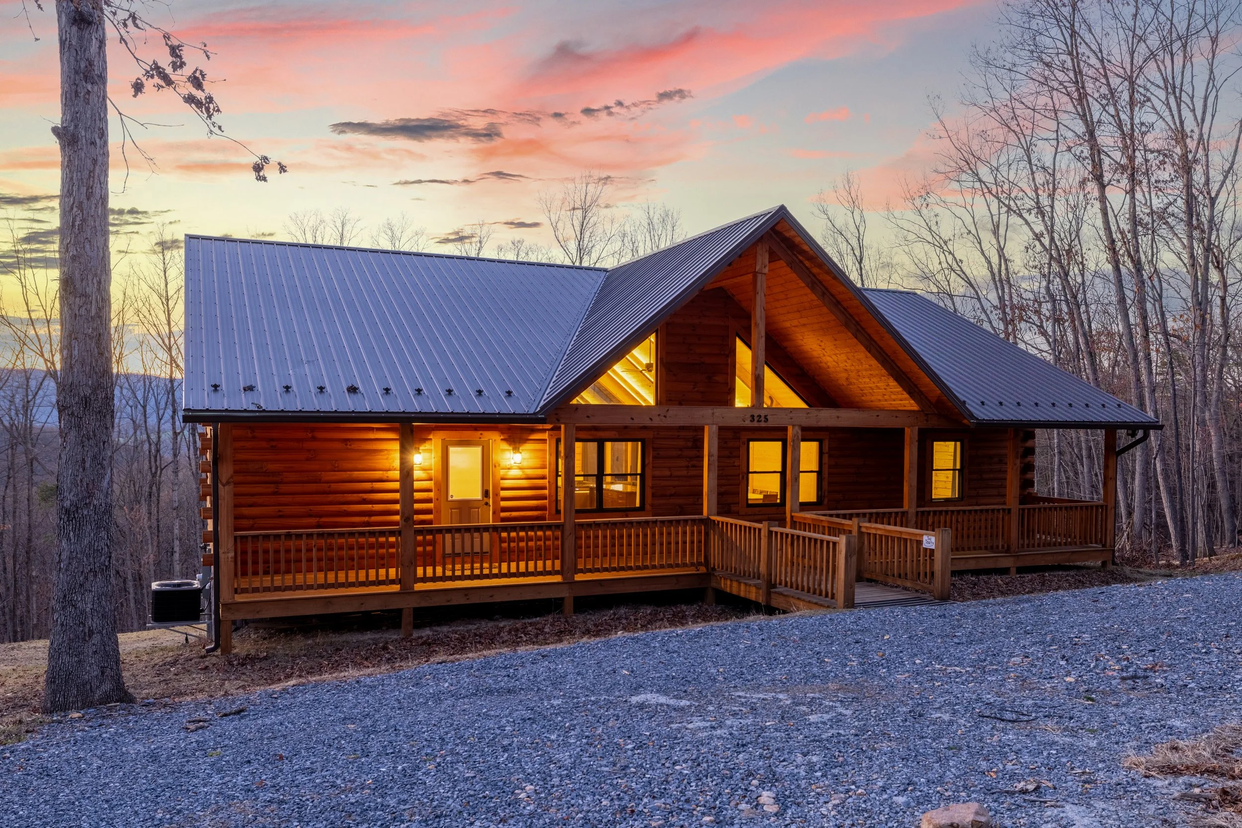 A wooden cabin with a metal roof is lit from inside during sunset, surrounded by leafless trees and a gravel path.