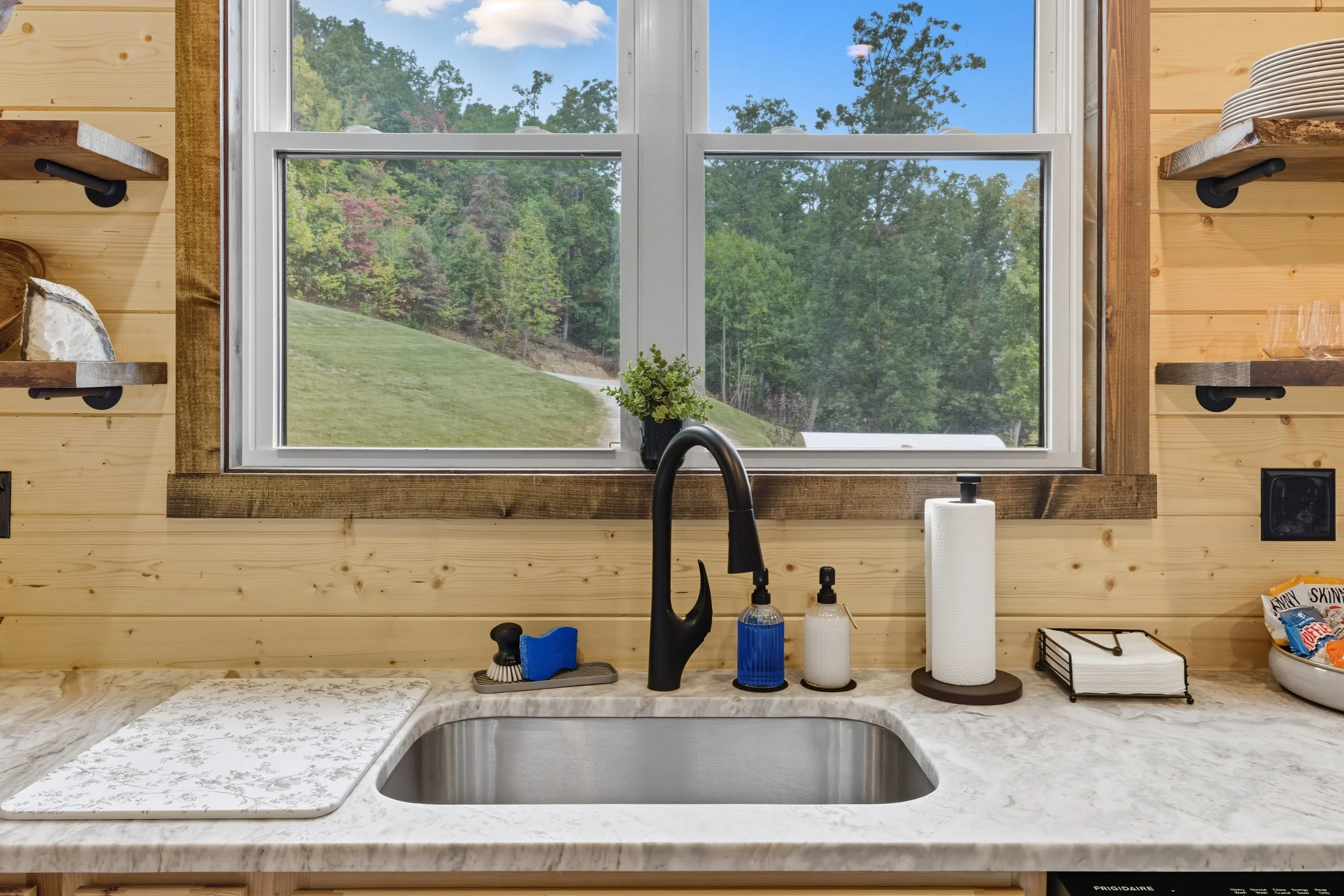 A kitchen window facing a grassy hill with trees in the distance, seen from the sink area with kitchen essentials on the counter.