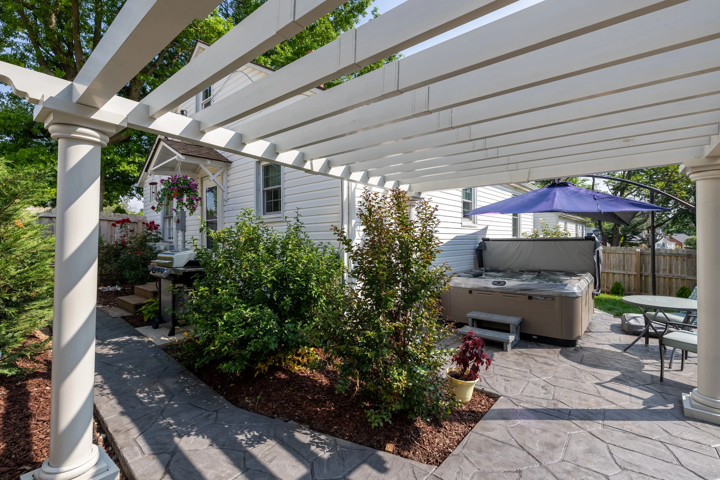 Backyard patio with white pergola, hot tub, patio furniture, and garden plants.