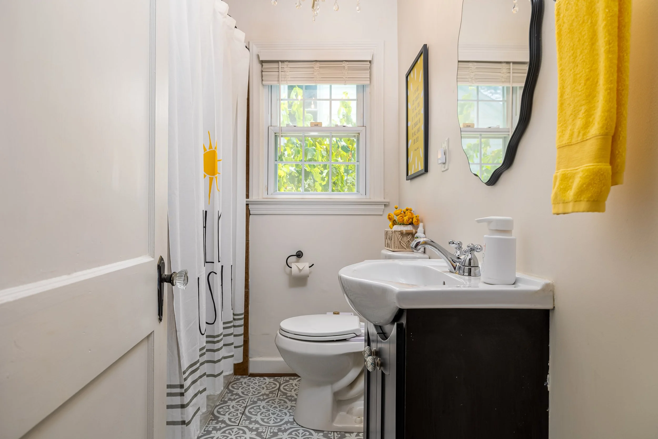 A small bathroom with a window showing green foliage outside. There is a white toilet, a sink with a chrome faucet, a soap dispenser, a small basket of yellow roses, a picture on the wall, a yellow towel, a shower curtain with a sun design, and decor