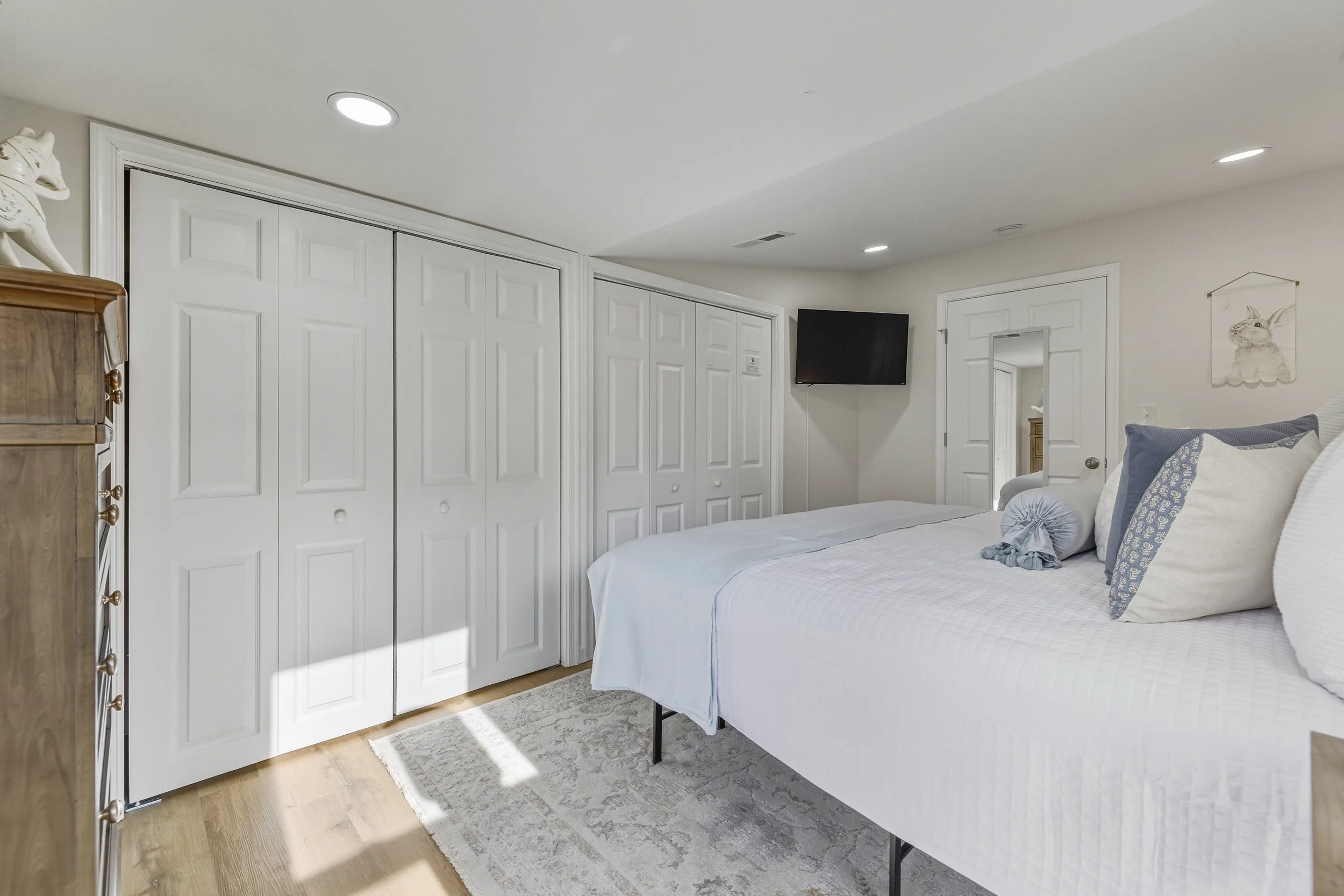 A bedroom with a white bed, pillows, and a small gray rug, white closet doors, a wall-mounted television, a wooden dresser, and wall art of a rabbit drawing.