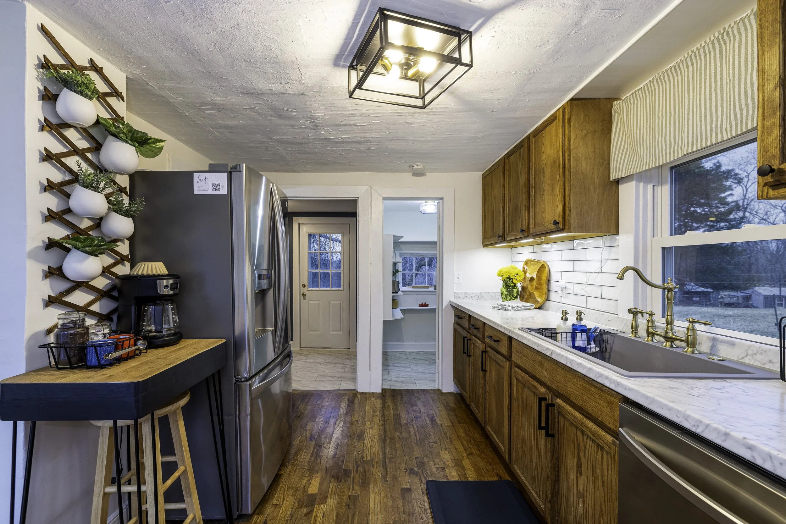 A cozy kitchen with wooden cabinets, a marble countertop, and a large window with a view of the outdoors. There are yellow flowers on the counter, a black dish rack, and a stainless steel refrigerator. The ceiling has a modern black square light fixture, and a wall-mounted wooden lattice with potted plants decorates one side.