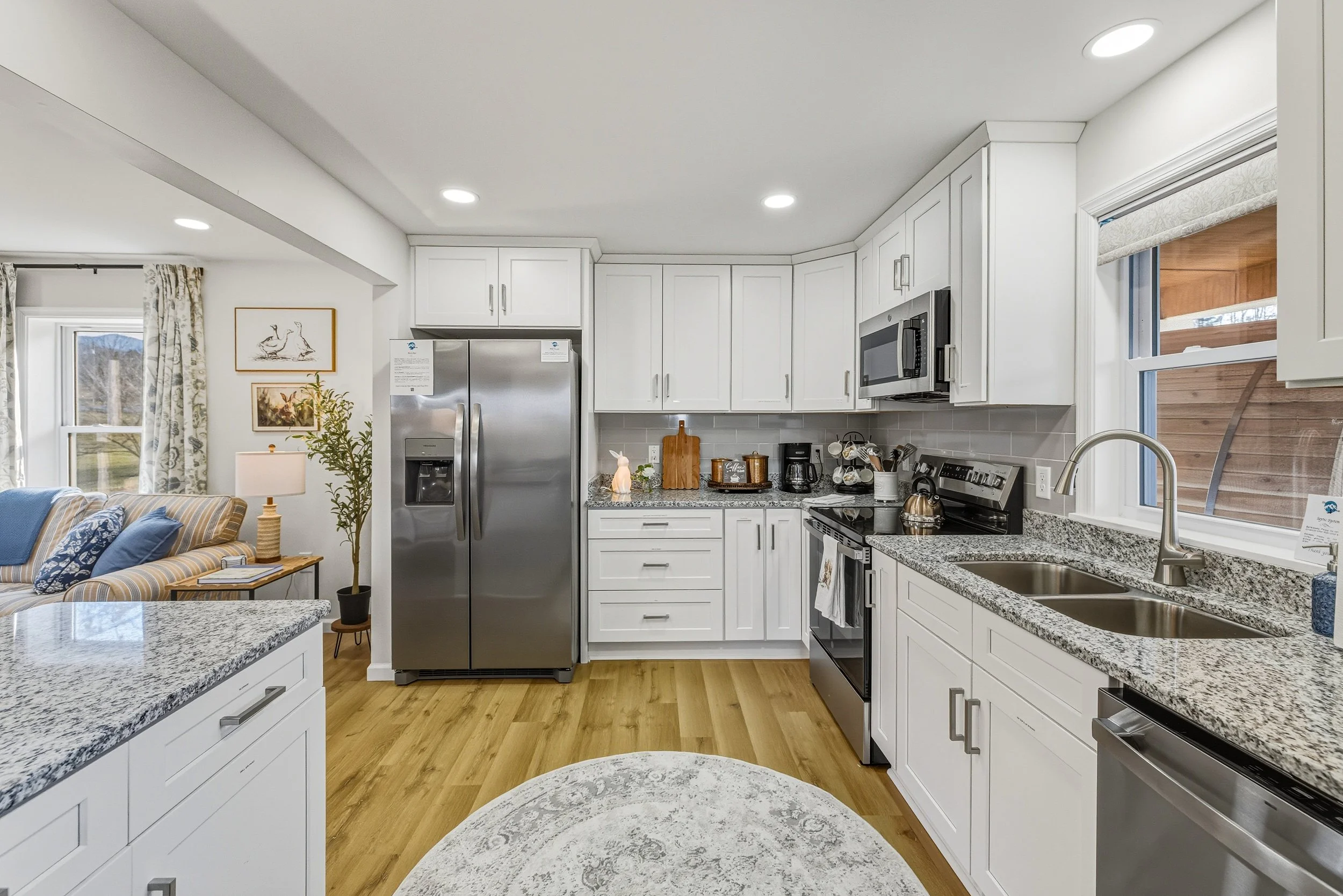Kitchen with white cabinets, stainless steel appliances, granite countertops, a window with a view of a wooden deck, and a living area with a striped sofa and artwork.