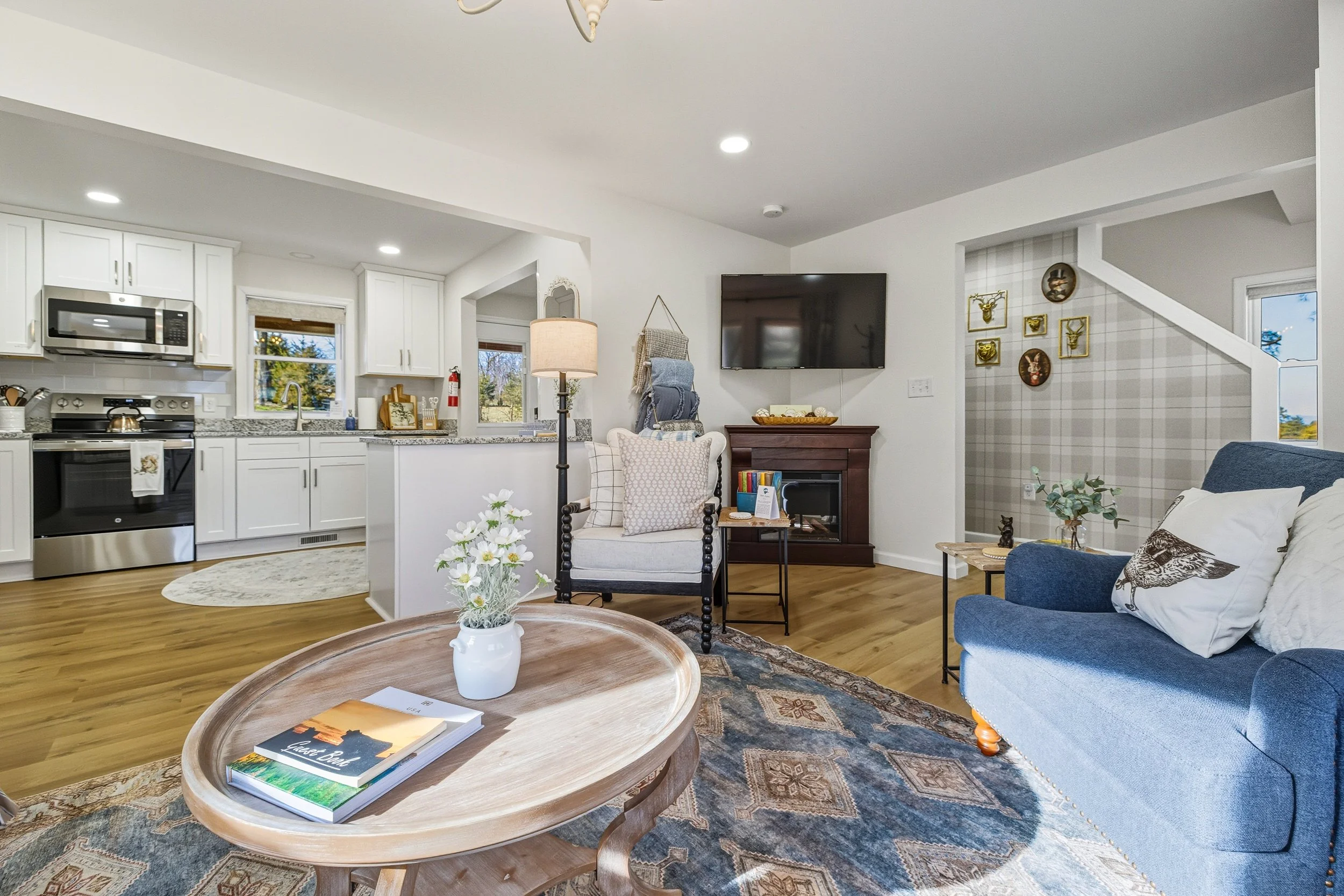 Living room with white kitchen cabinets, stainless steel stove, microwave, and granite countertops, a flat-screen TV on the wall, a comfortable blue sofa with pillows, a wooden coffee table with books and a white vase with flowers, and decorative wal