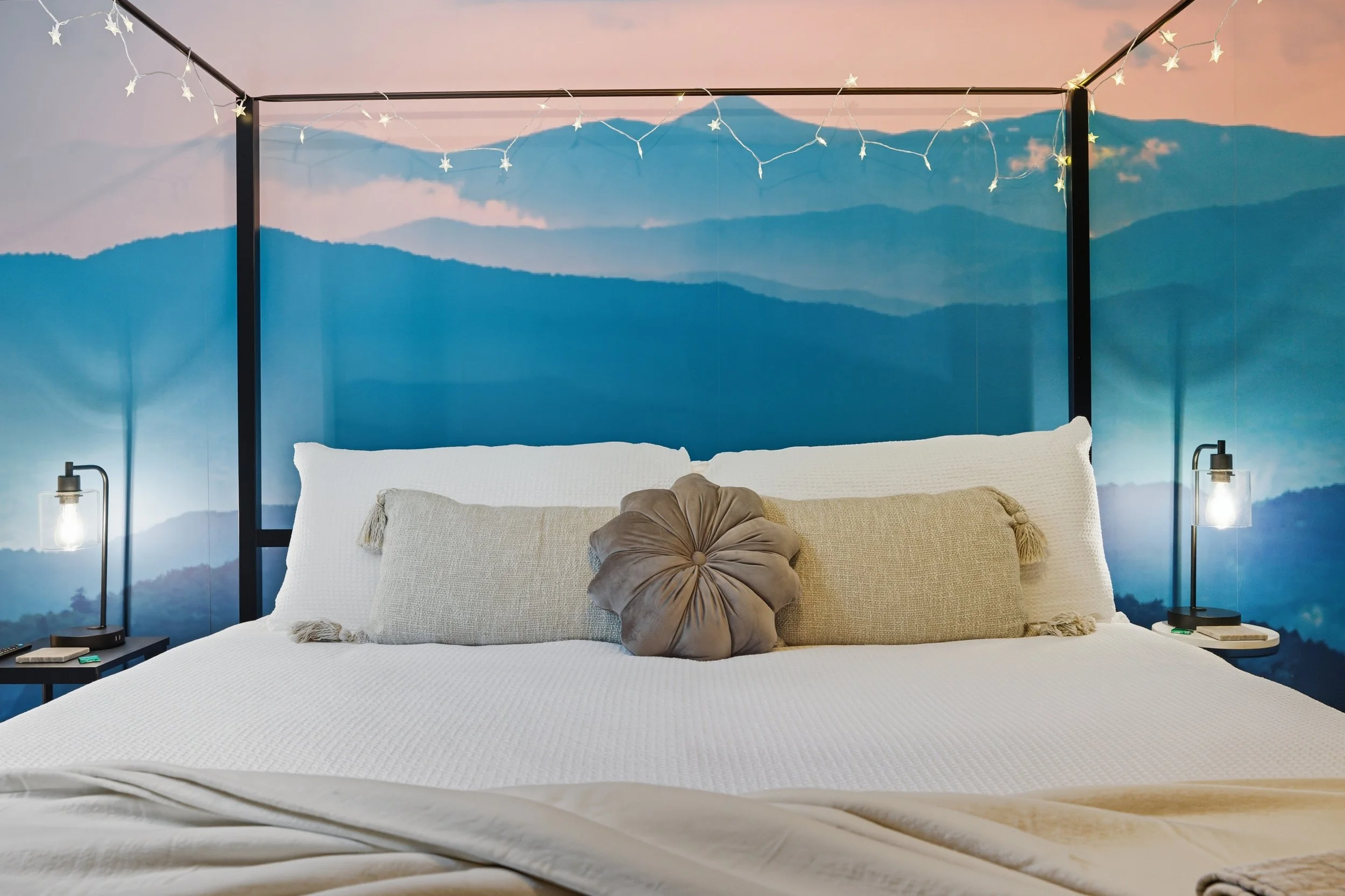A bedroom with a mountaintain mural on the wall, string lights hanging from the ceiling, a white bed with multiple pillows, two black bedside tables with lamps, one of which has a book and a music box on it.