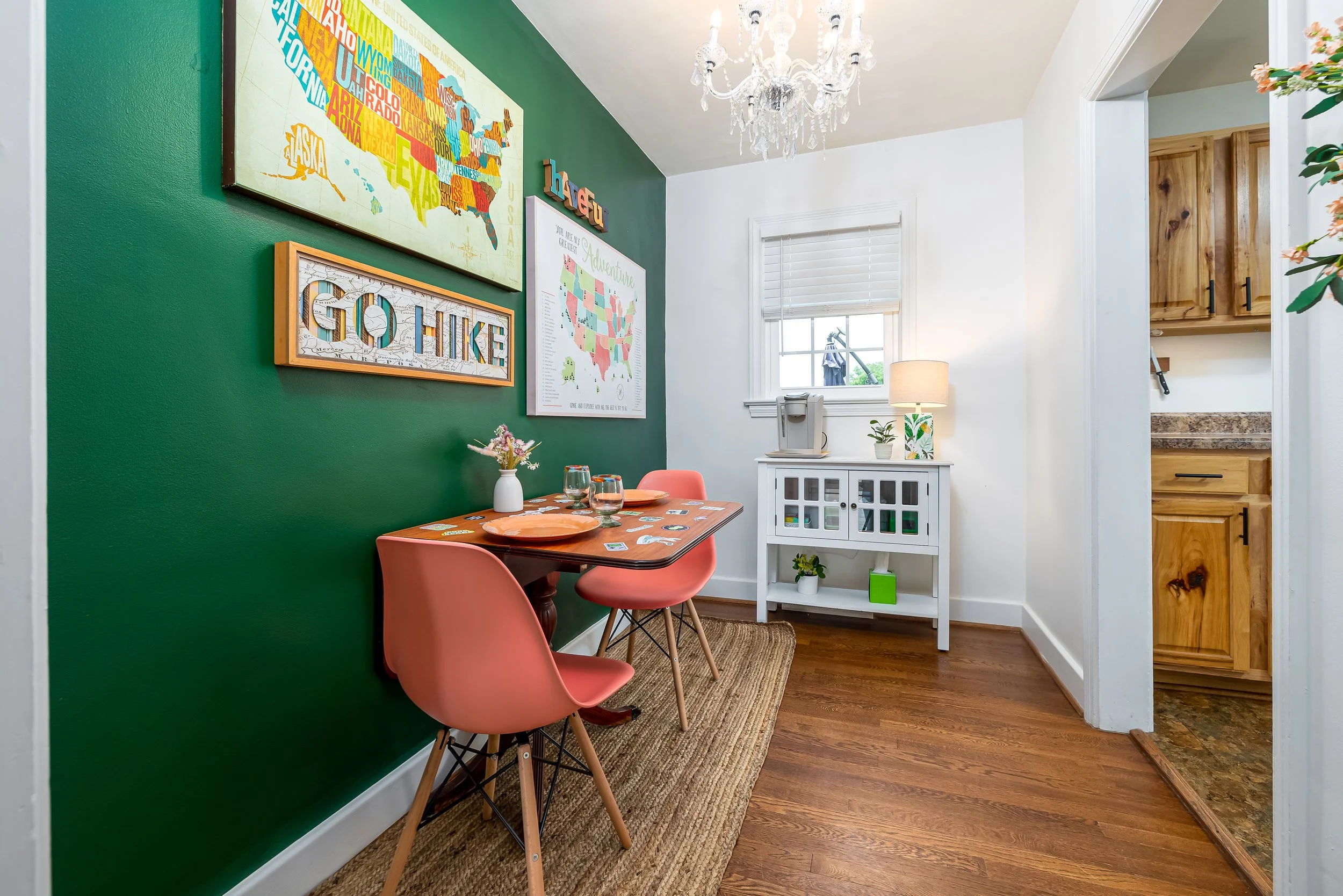 The image shows a dining area with a green accent wall decorated with colorful maps and signs, including a large map of the United States, a 'GATHER' sign, and a chalkboard with the word 'SMOKE'. The dining table is set with orange plates, glasses, a