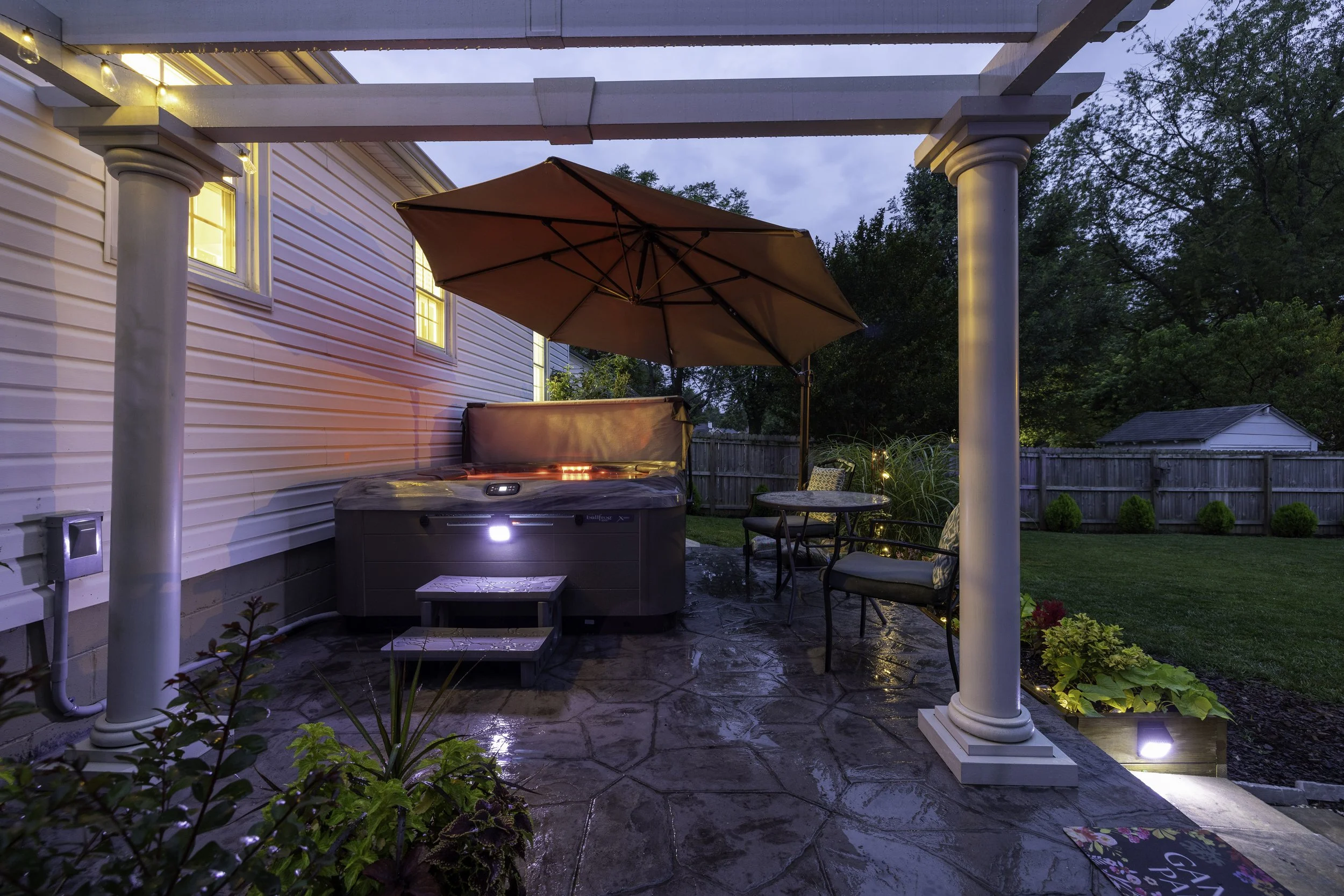 A backyard patio area with a covered space featuring two columns, with an orange patio umbrella over a hot tub, and outdoor furniture illuminated by string lights and landscape lighting at dusk.