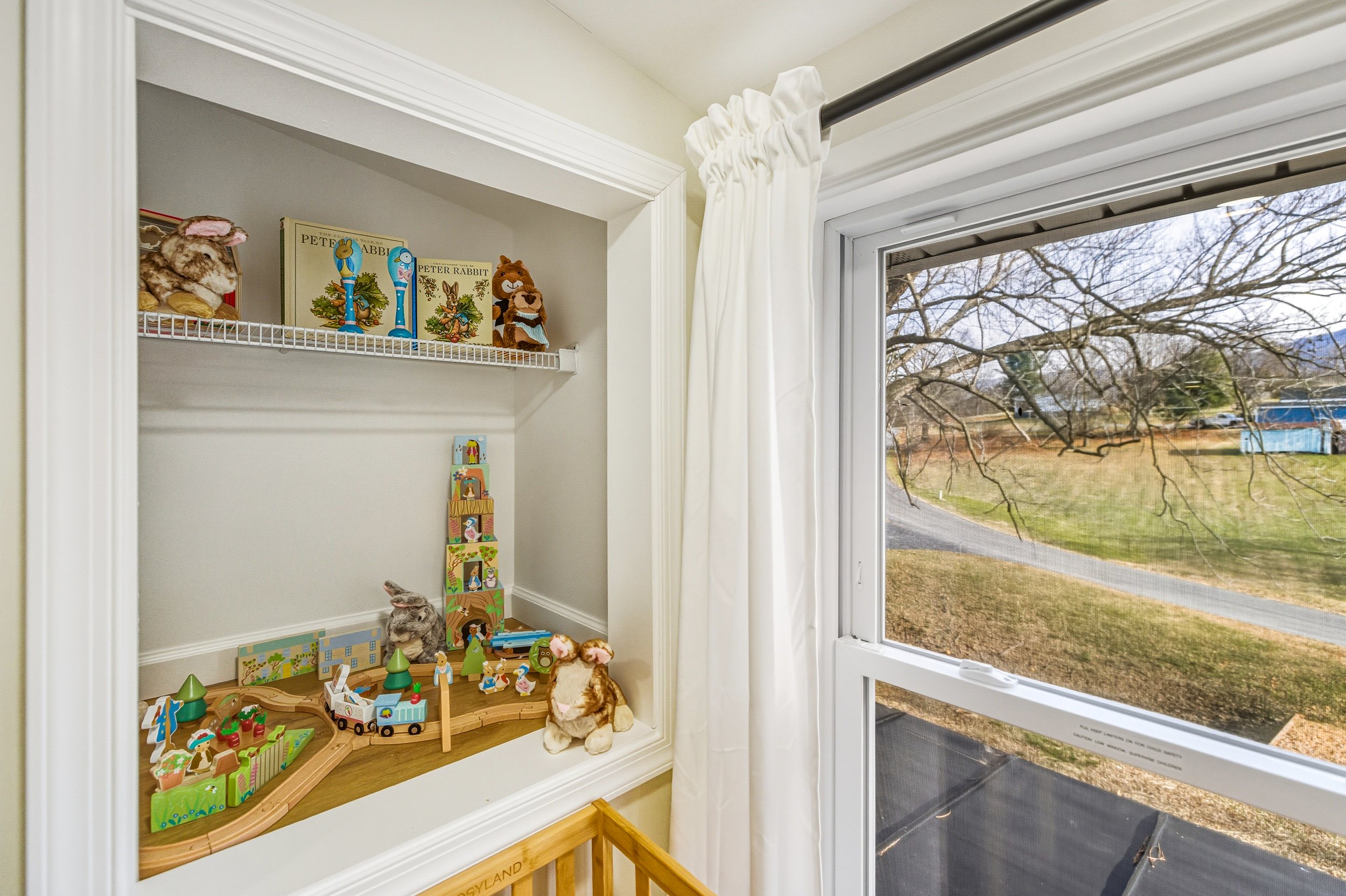A collection of children's toys and plush animals on a white built-in window seat, with a toy train and wooden blocks, next to a window with white curtains and a view of leafless trees outside.