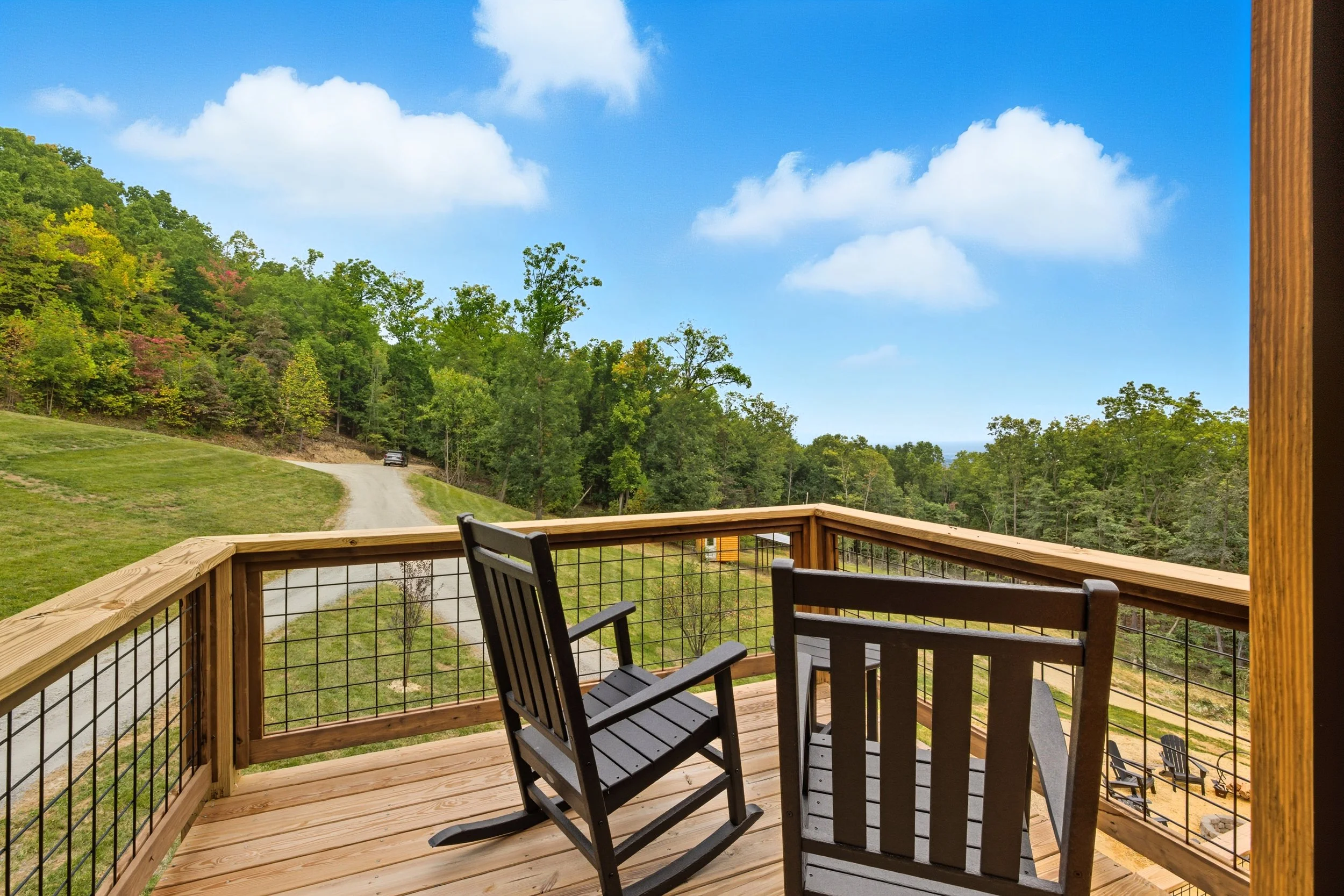A wooden balcony with black chairs overlooking a green landscape with trees and a dirt road, under a blue sky with scattered clouds.