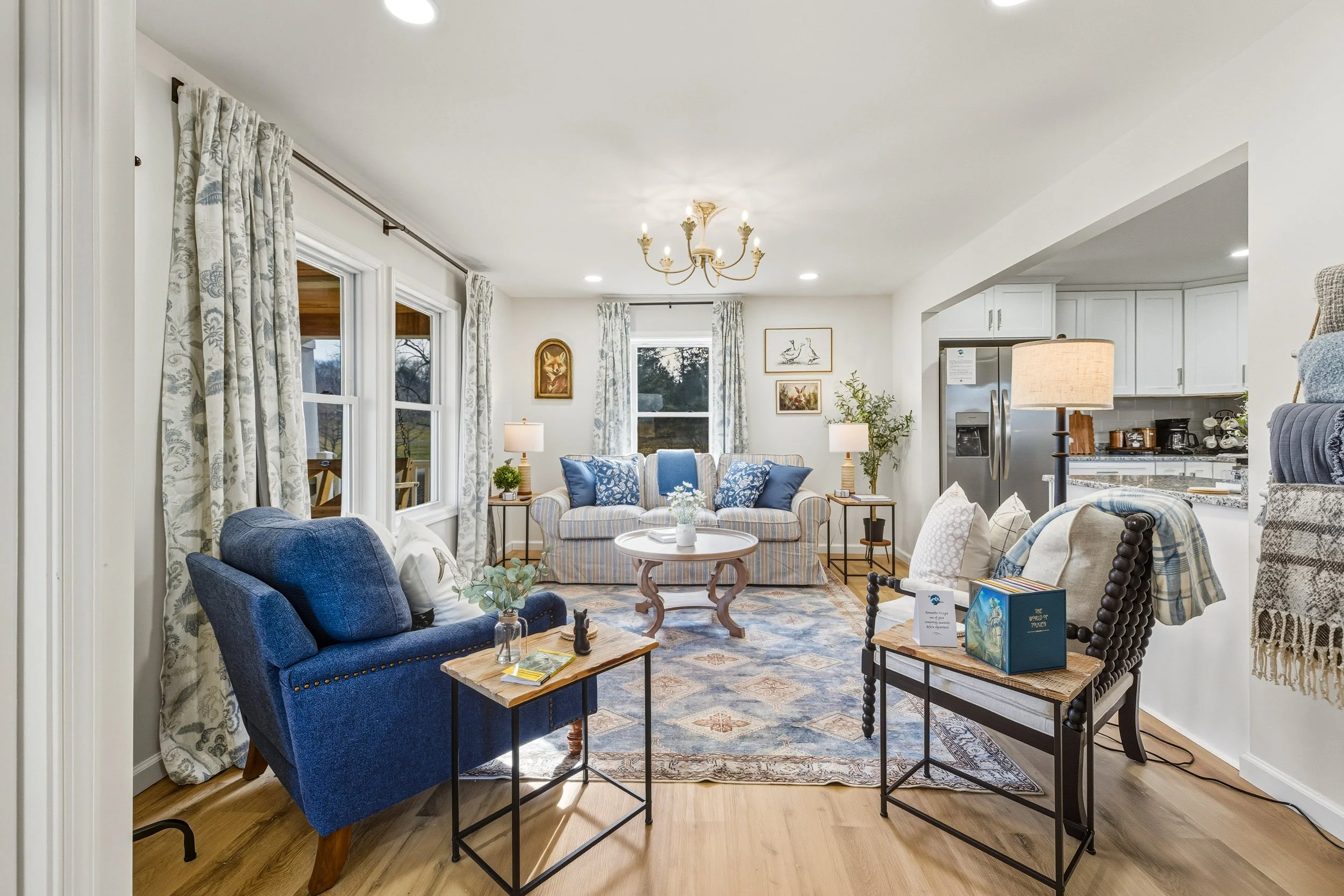 Living room with blue and striped upholstered furniture, a chandelier, framed artwork, a kitchen opening, and numerous windows with floral curtains.