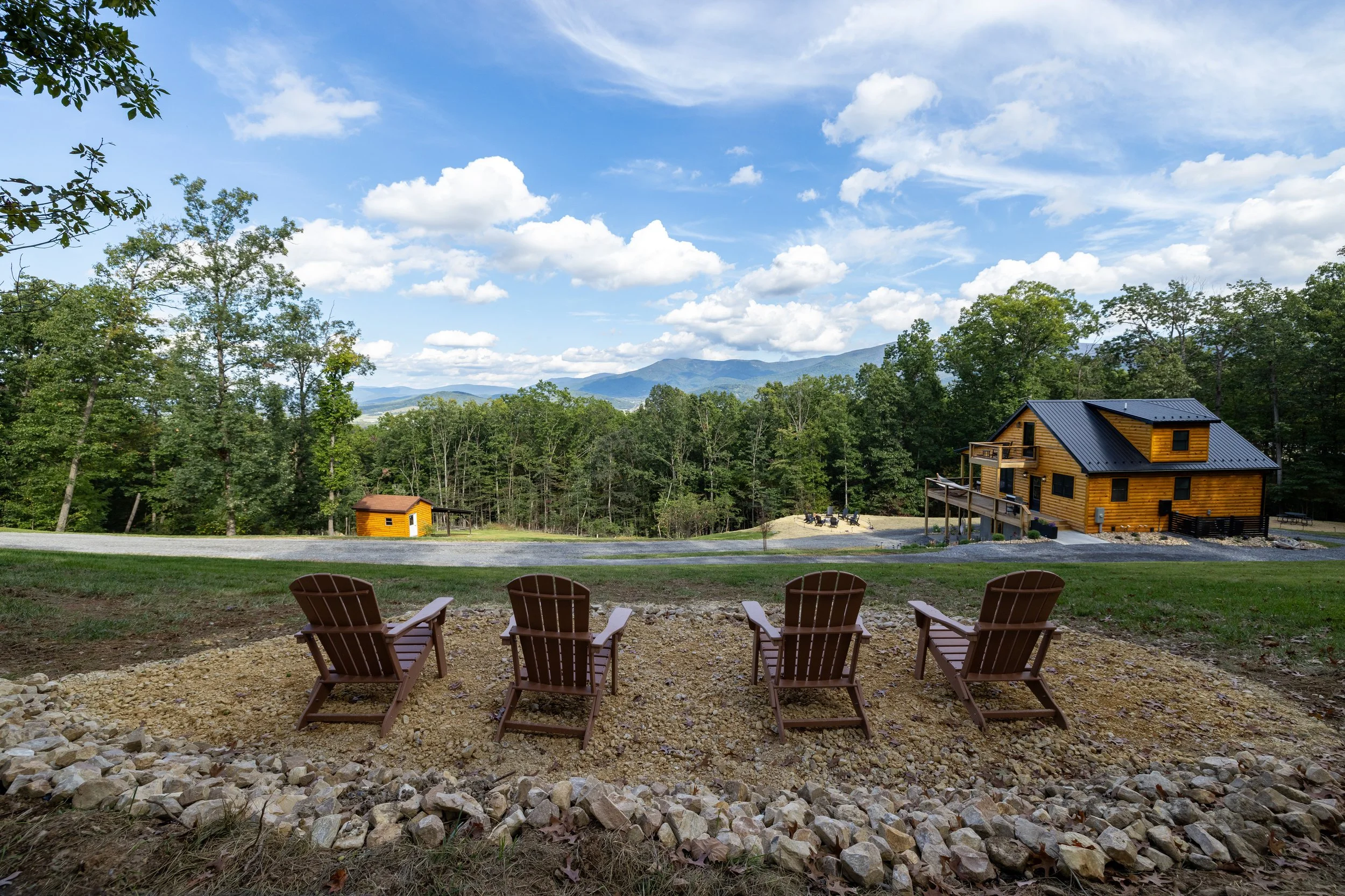A scenic outdoor view of a mountain landscape with a blue sky and clouds, a wooden house with a metal roof, green trees, a gravel path, and four brown wooden chairs in the foreground.