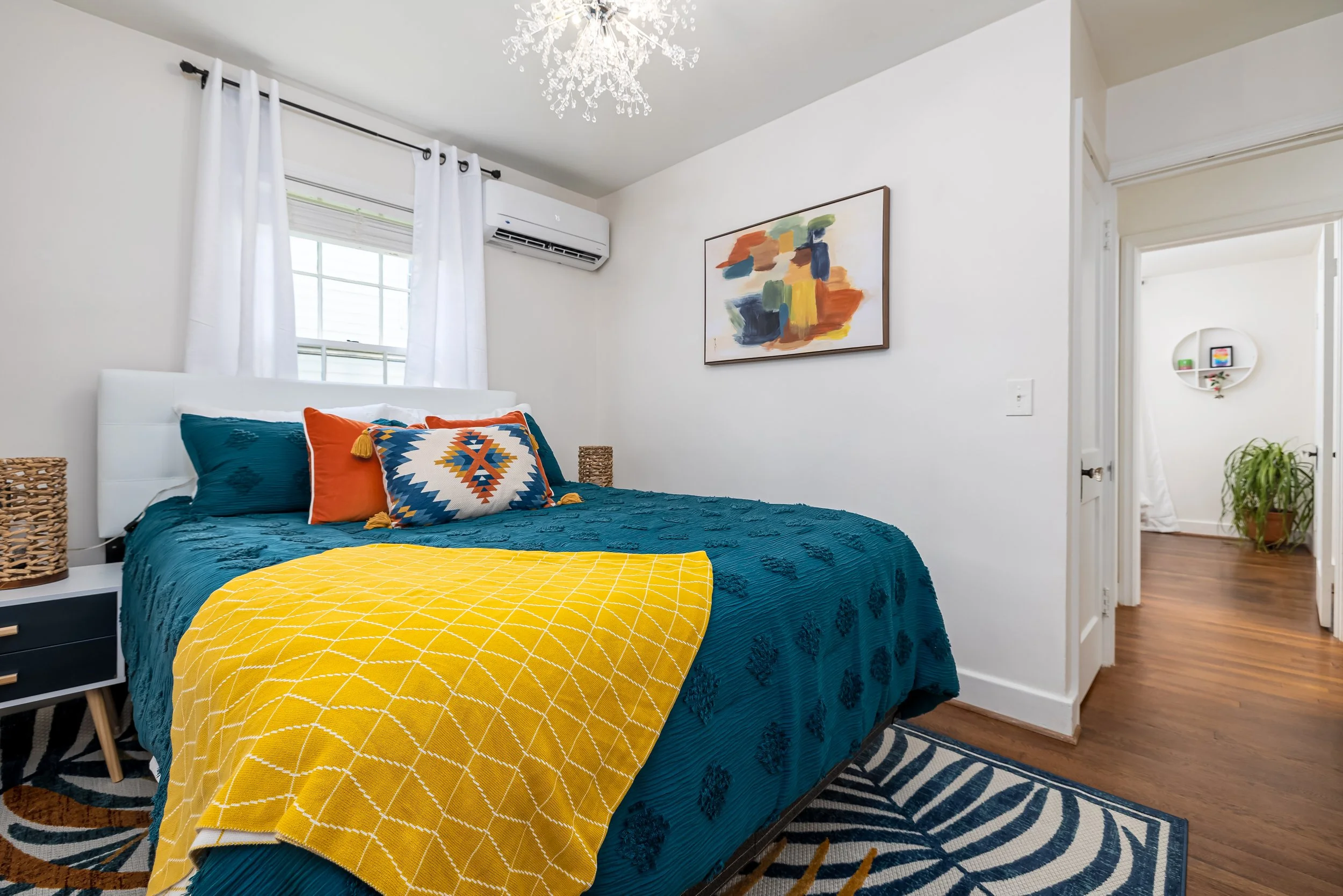 A bedroom with a blue bedspread, orange and patterned pillows, a yellow blanket, a white headboard, a side table, a window with white curtains, wall art, an air conditioner, and a patterned rug, with a doorway leading to another room with a circular 