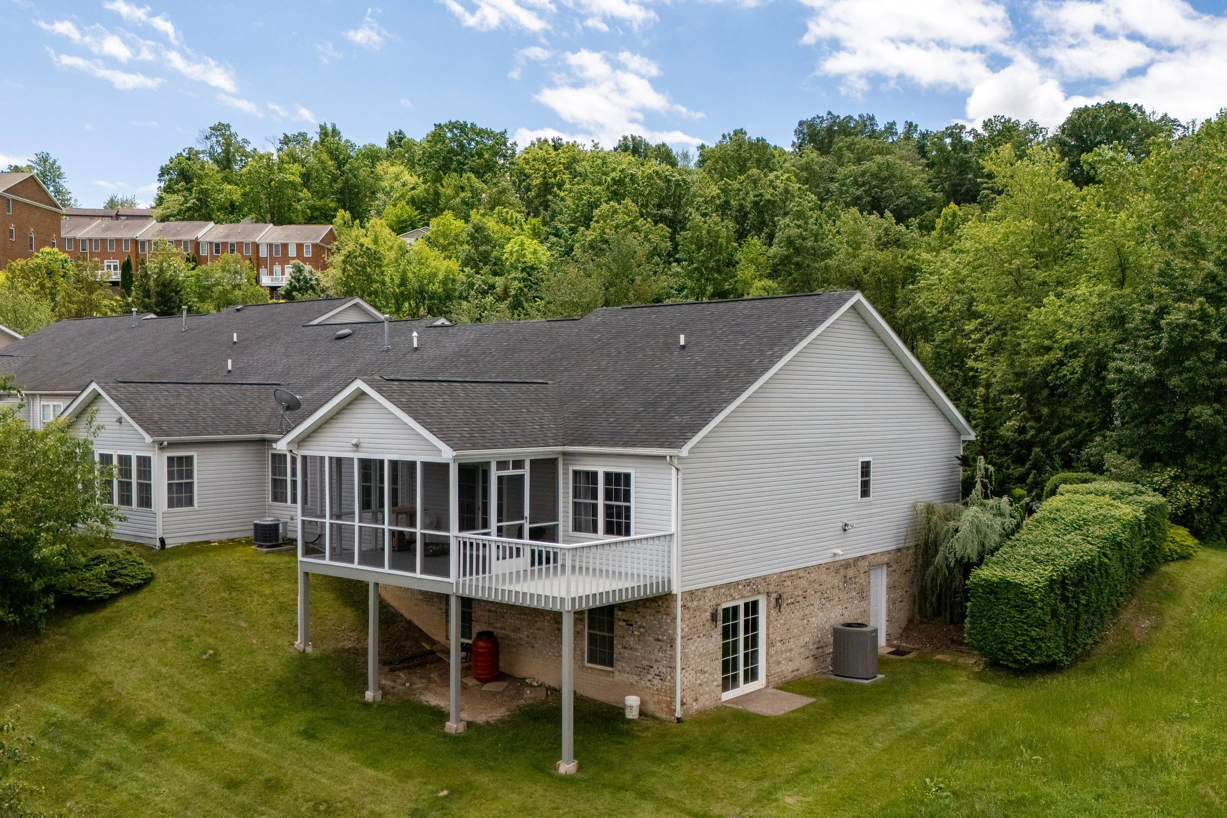 A suburban backyard with a white house featuring a screened porch, surrounded by green grass and trees, with neighboring houses visible on a hill in the background under a partly cloudy sky.