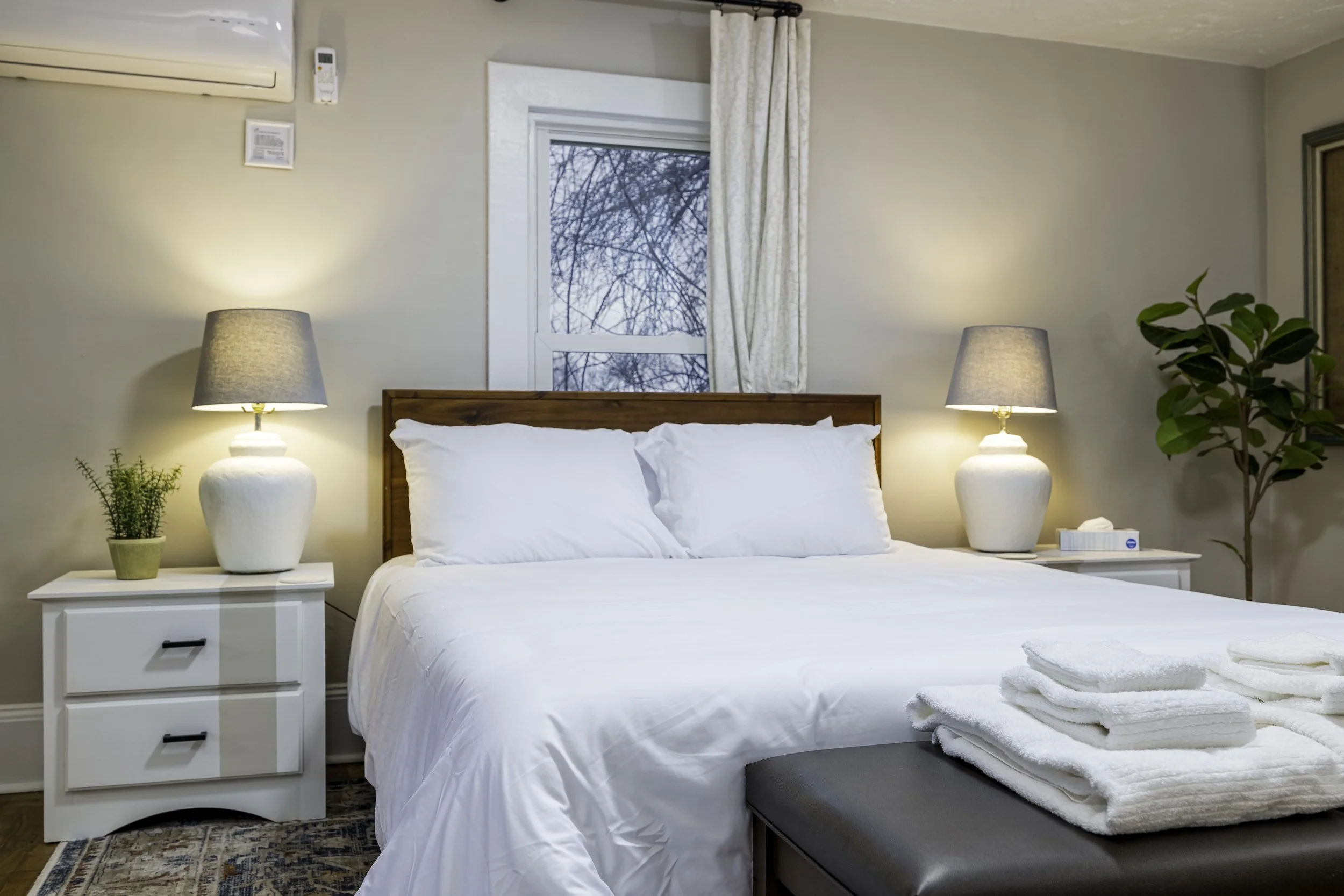 A neatly made bed with white bedding and pillows in a bedroom featuring two matching bedside tables with lamps, a window with curtains, a potted plant, and a stack of folded towels at the foot of the bed.