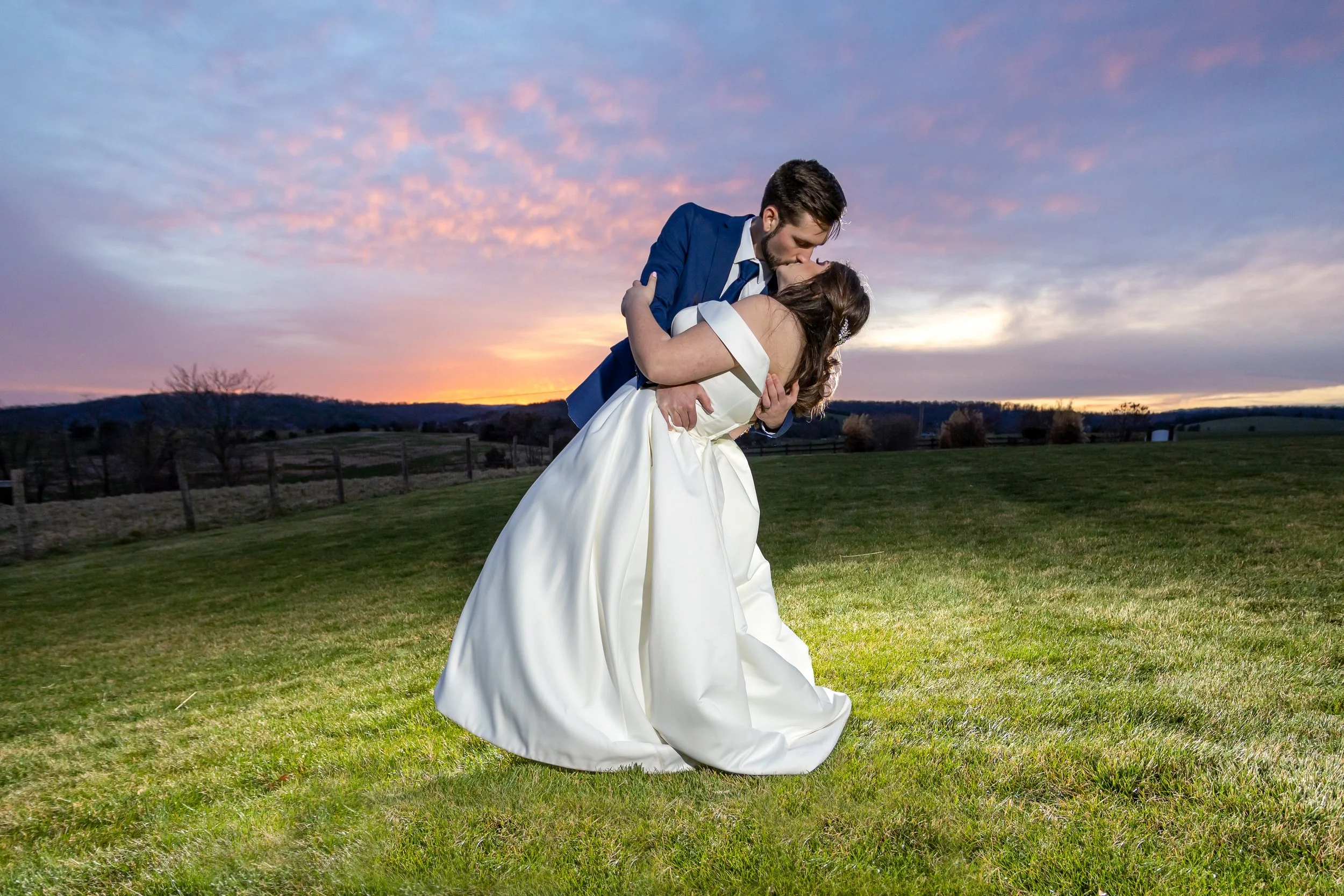 A bride and groom sharing a kiss outdoors at sunset, with the groom dipping the bride, both dressed in wedding attire, on a grassy field with a scenic landscape and colorful sky in the background.
