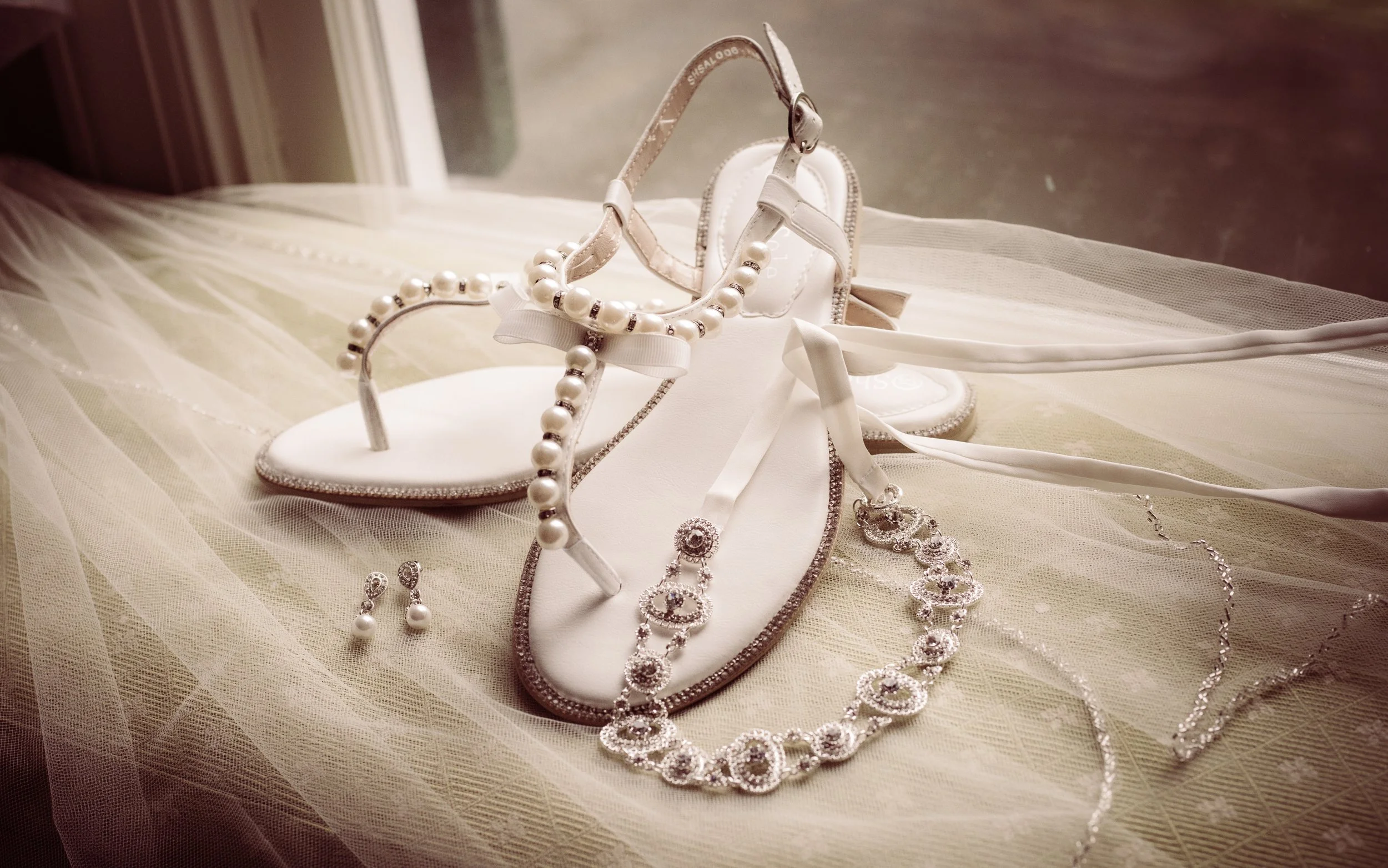 Bridal sandals with pearl embellishments, a pearl necklace, pearl earrings, and white lace fabric.